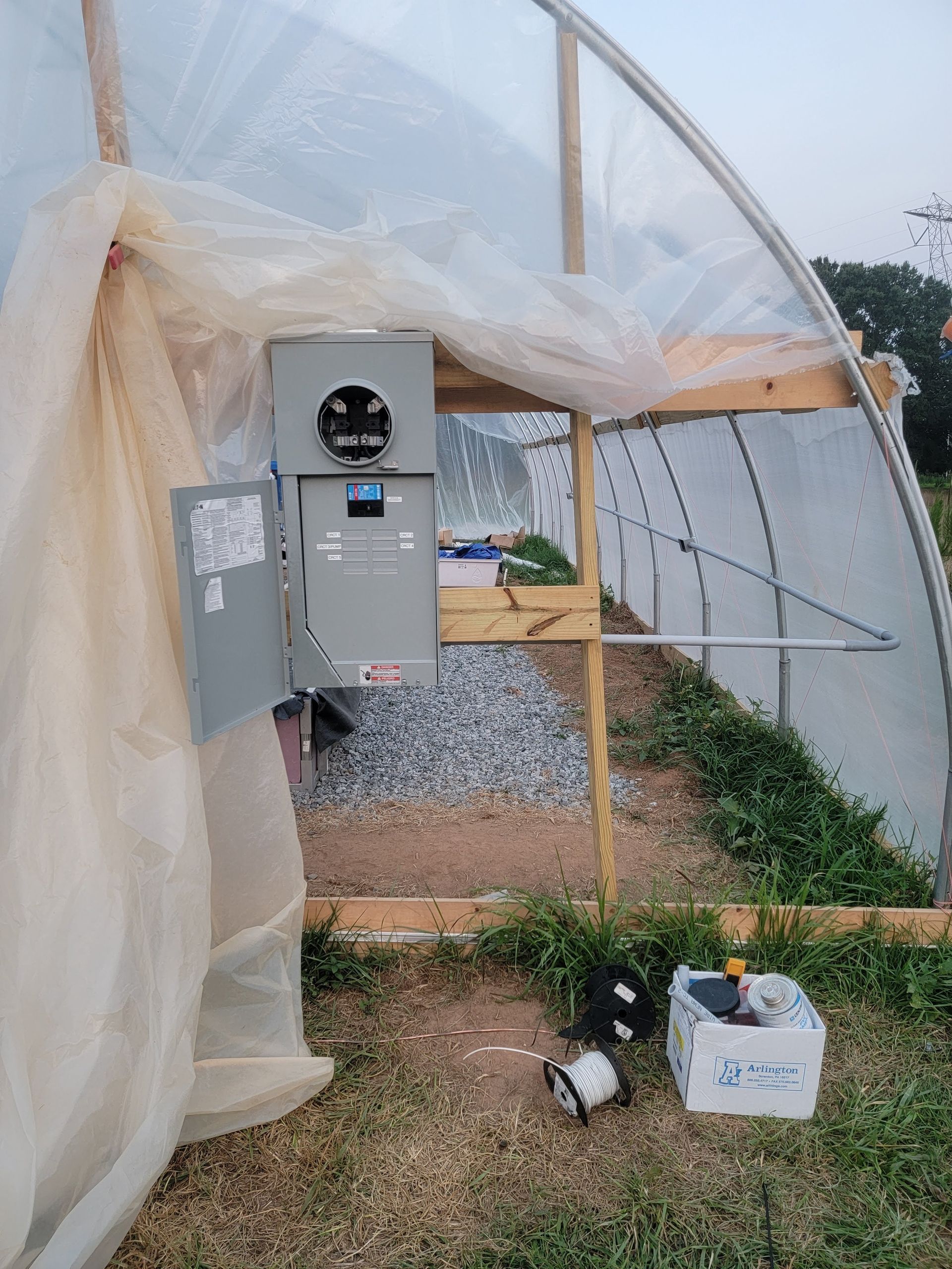 Plastic-covered greenhouse with an electrical control panel and supplies inside a dirt-floored entrance