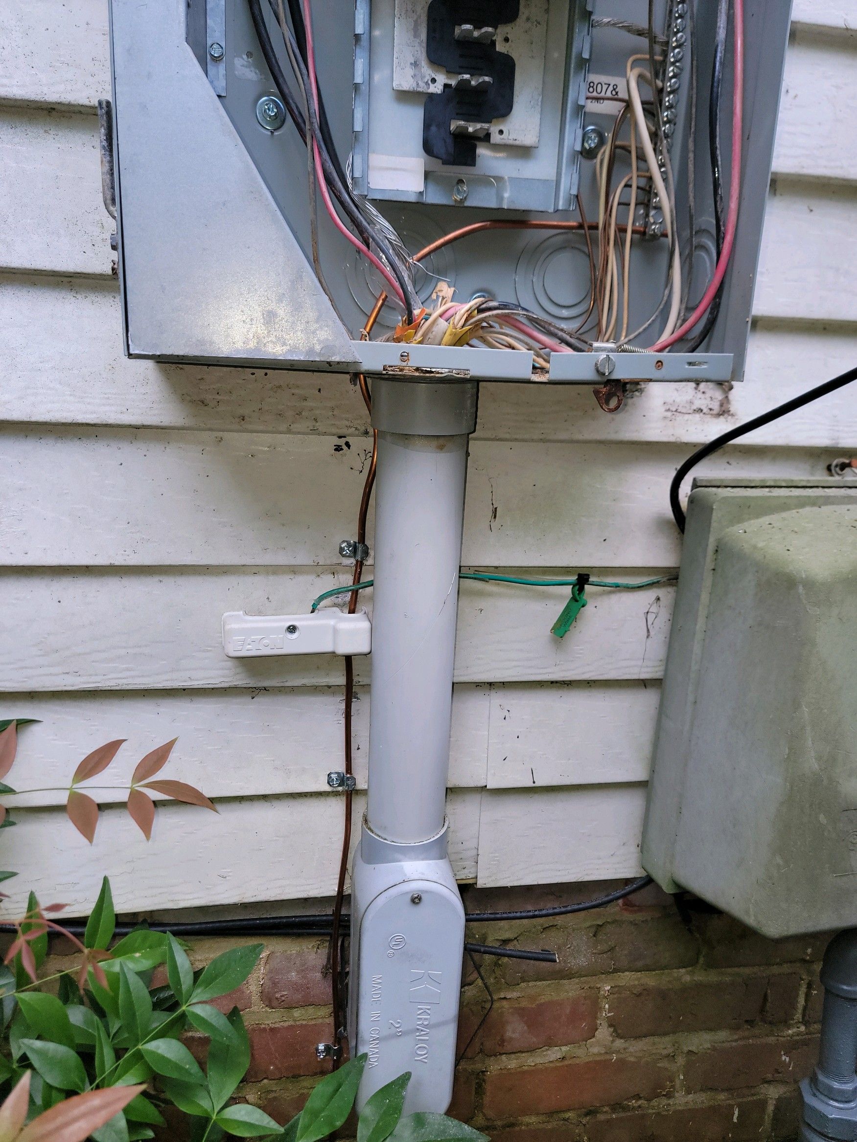 Open electrical panel with exposed wires above a gray conduit on an exterior house wall