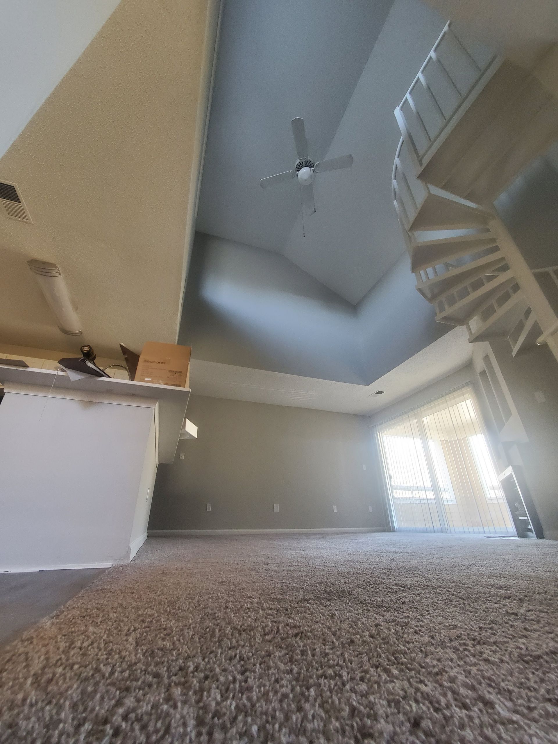 Empty carpeted living room with staircase, vaulted ceiling, and bright window blinds