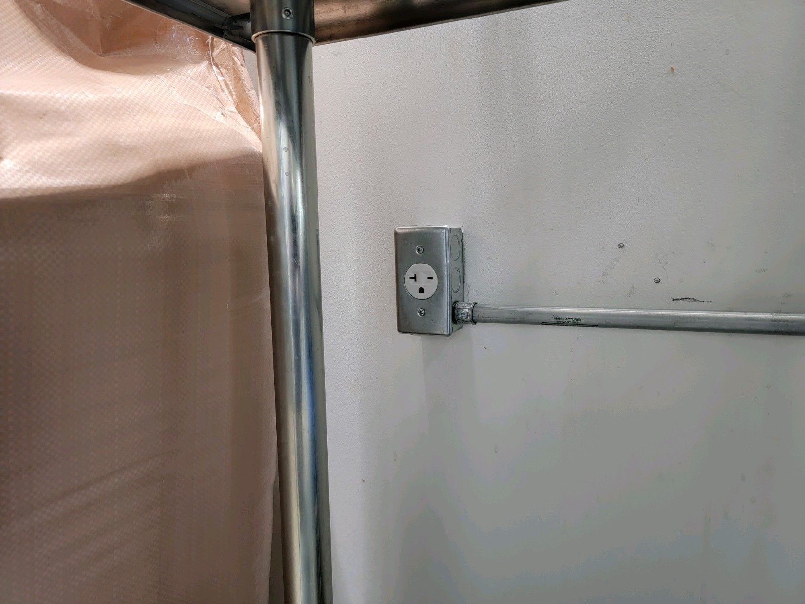 Wall corner with exposed electrical outlet, conduit, and a pink curtain or partition on the left.