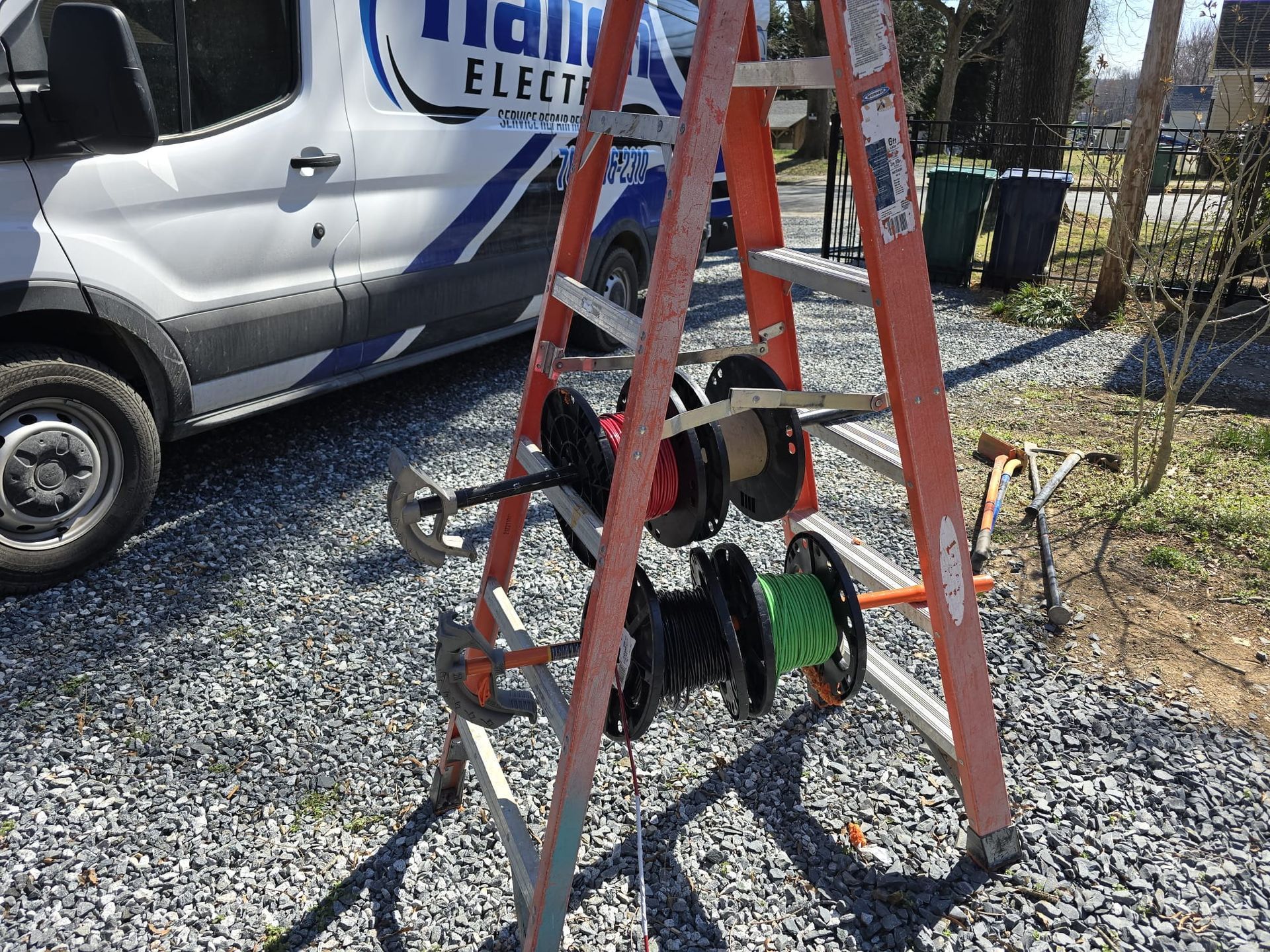 Orange ladder holding a spool of black cable beside a white utility van on gravel