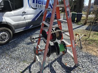 Orange ladder holding several cable reels outdoors beside a white work van on gravel.