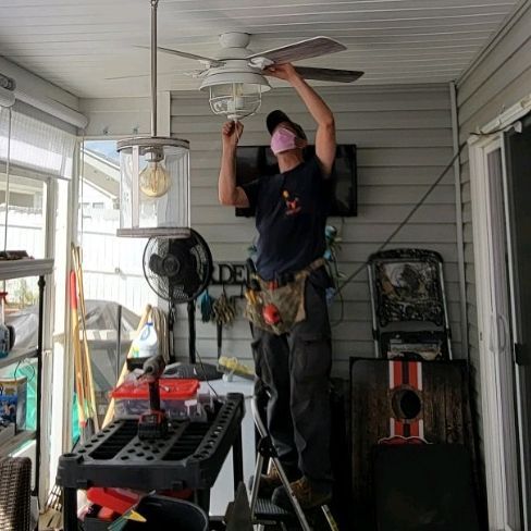 Person on a porch adjusting a ceiling fan light with tools nearby.