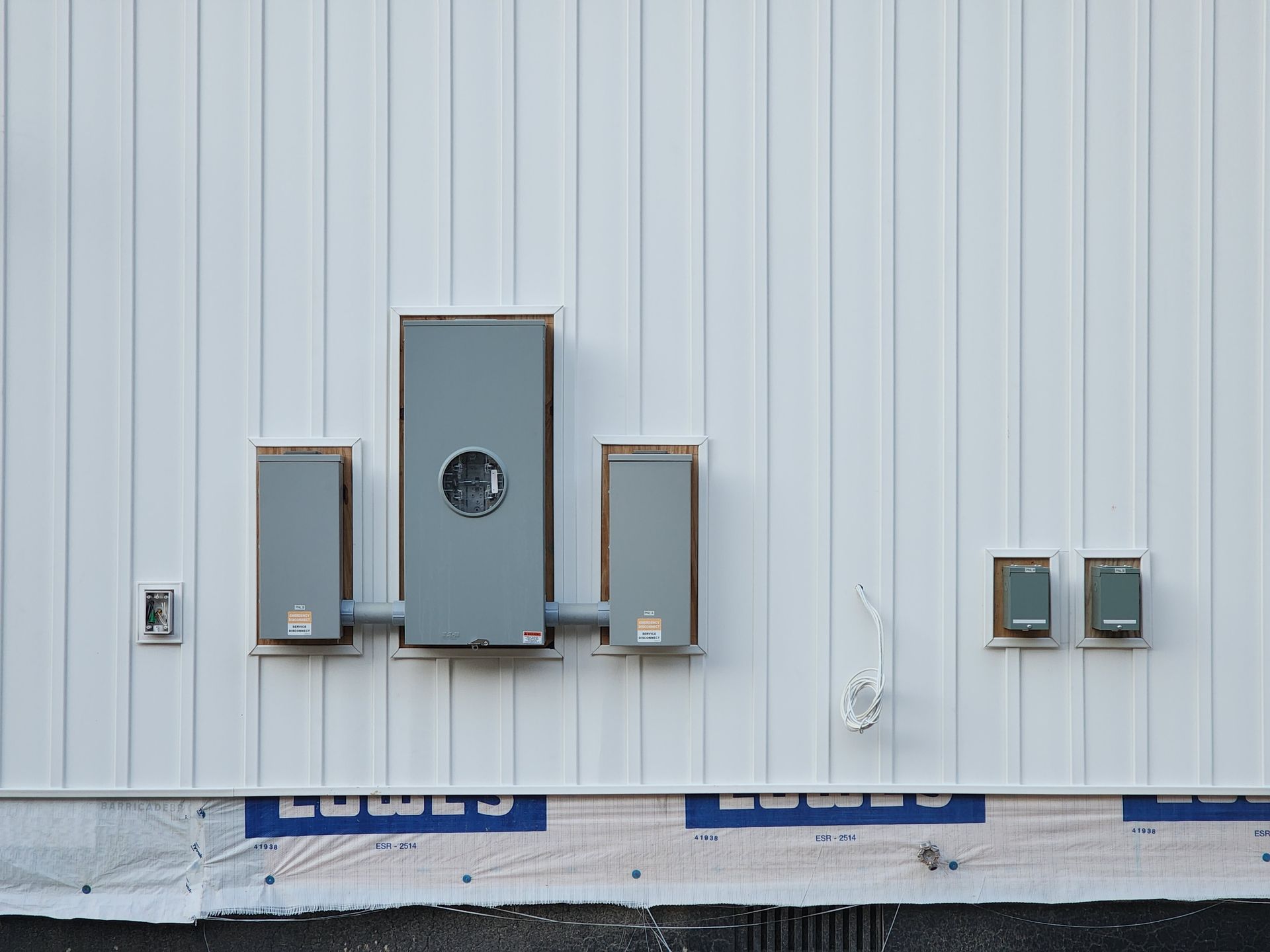 Electrical meter boxes mounted on a white exterior wall near a partially covered foundation