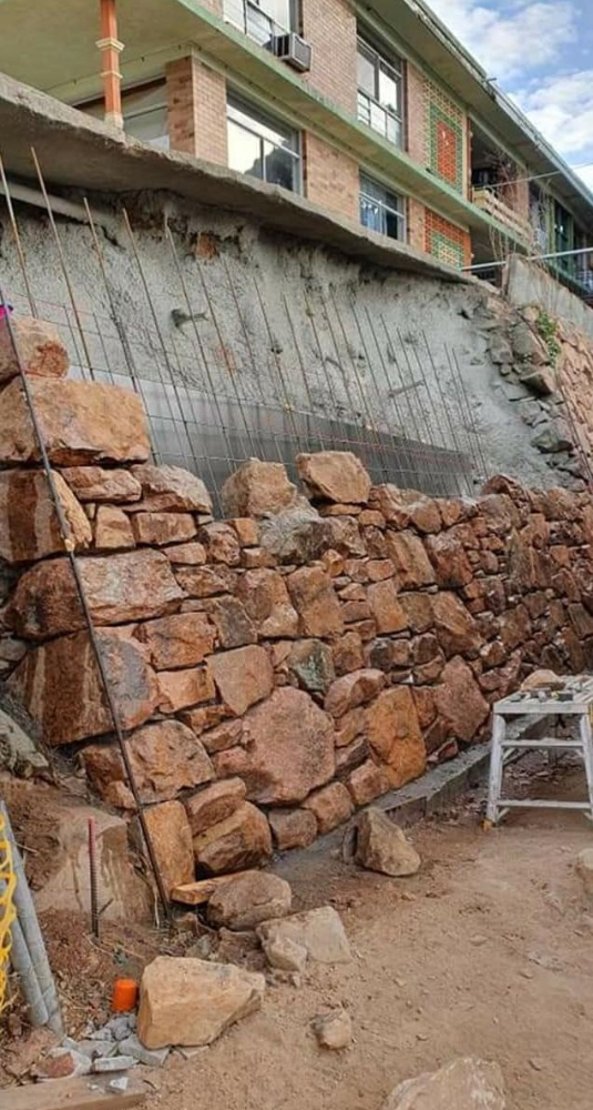 Stone retaining wall under construction beside a building on a dirt slope, with scaffolding visible above— High End Stone Masons in West End, QLD