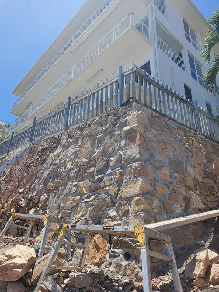 Stone retaining wall under construction beside a white building with metal railing and scaffolding.— High End Stone Masons in West End, QLD