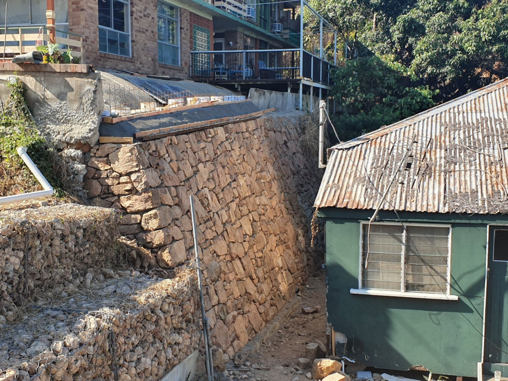 Steep stone retaining wall beside hillside houses, with a green building and corrugated metal roof in front— High End Stone Masons in West End, QLD