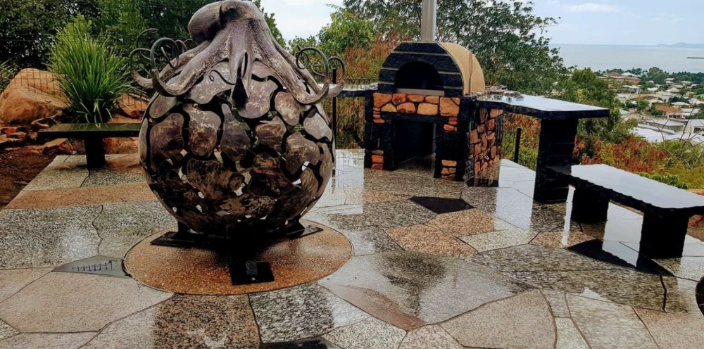Stone outdoor seating area with a sculptural fire pit, wet tiled floor, and ocean view in the background— High End Stone Masons in West End, QLD