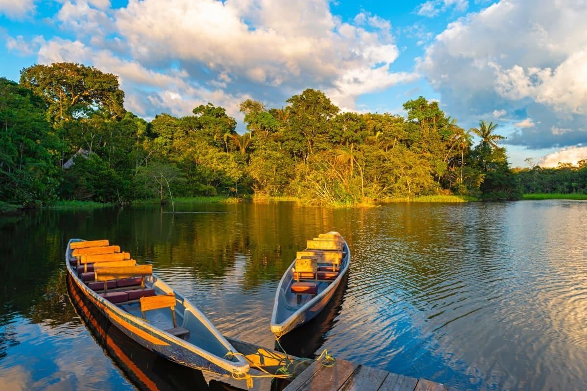 Dos barcos de madera en un río tranquilo al atardecer, con frondosos árboles verdes y un cielo nublado reflejado en el agua.