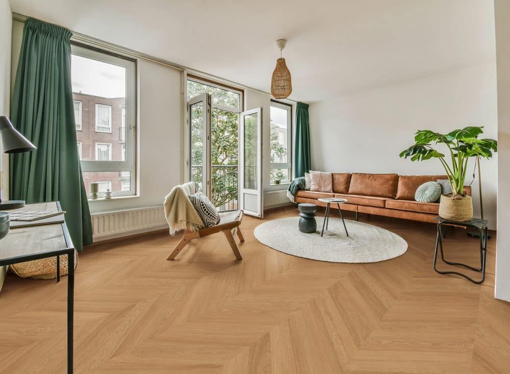 Helles Wohnzimmer mit beigefarbenem Sofa, rundem Teppich, großen Fenstern, offener Tür und Fischgrätparkett