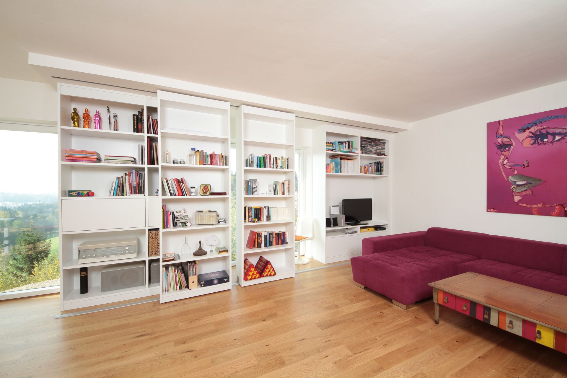 Helles Wohnzimmer mit weißen Bücherregalen, Holzboden und einem lila Ecksofa