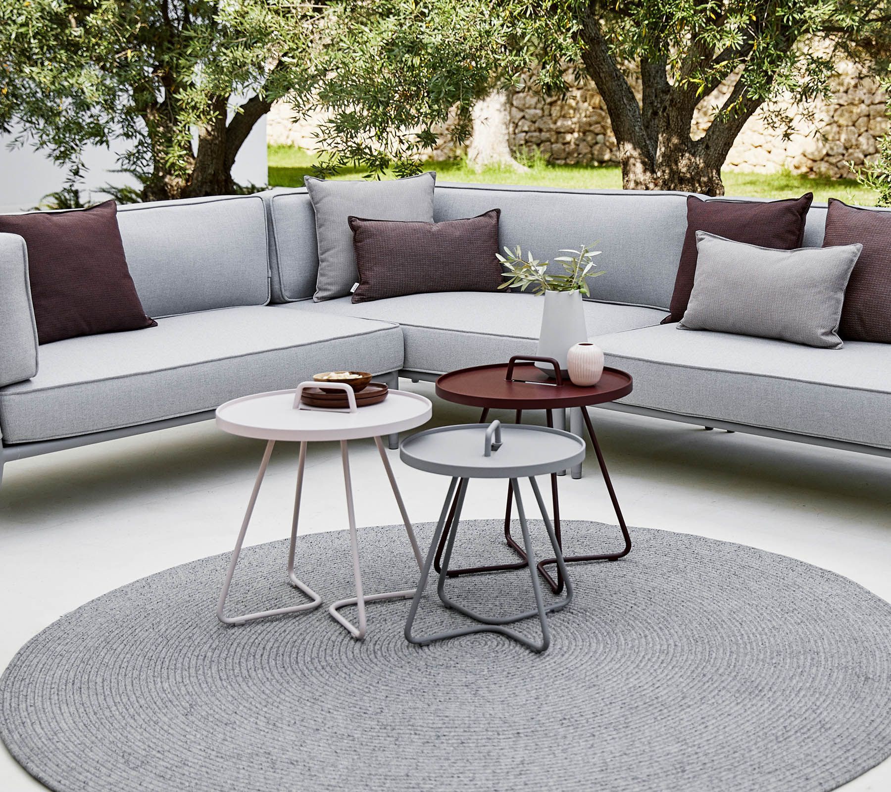 Terrasse im Freien mit grauem Ecksofa, runden Beistelltischen und rundem grauen Teppich unter Bäumen