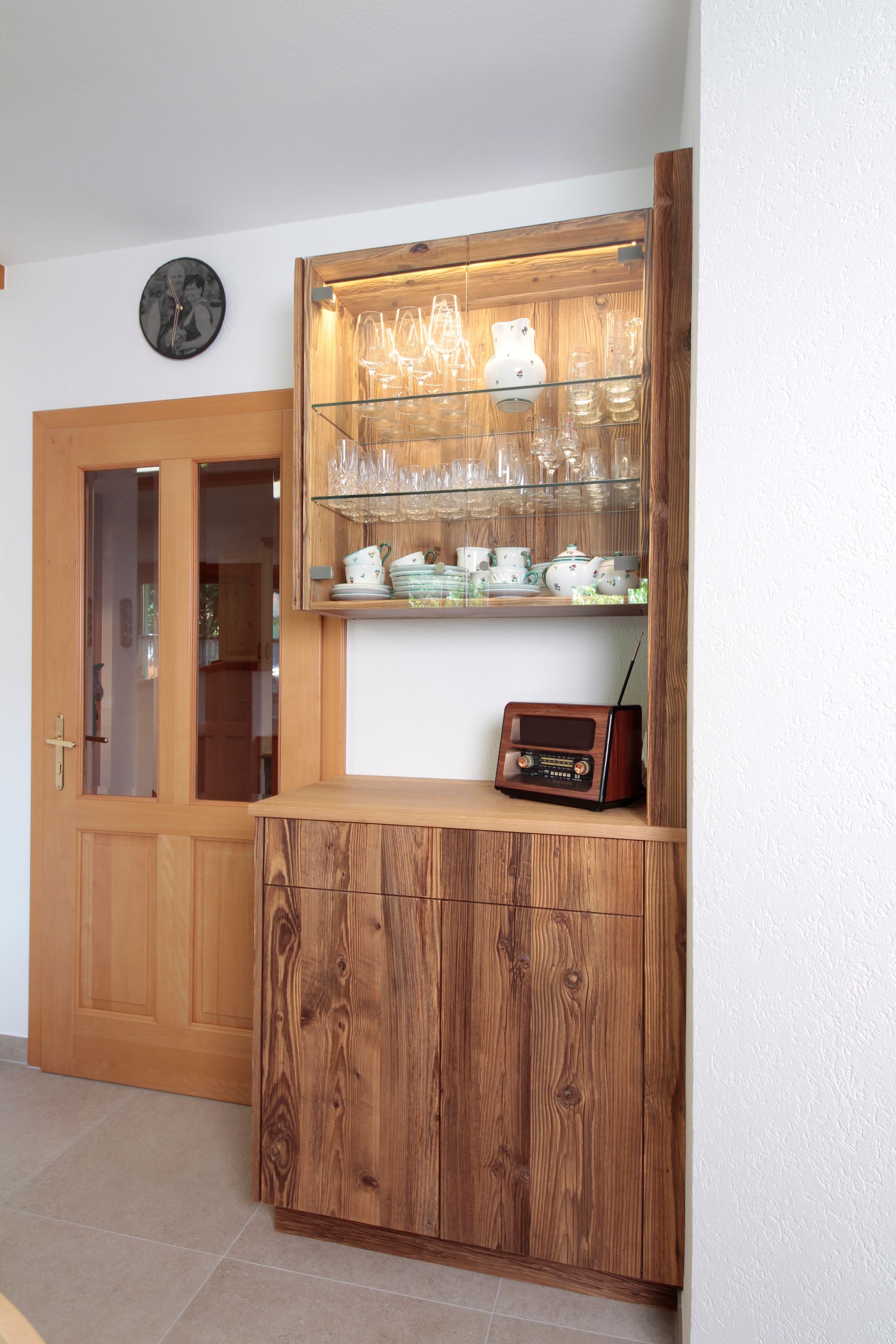 Ein Holzschrank mit Glasvitrinen steht in einem hellen Raum neben einer Holztür.