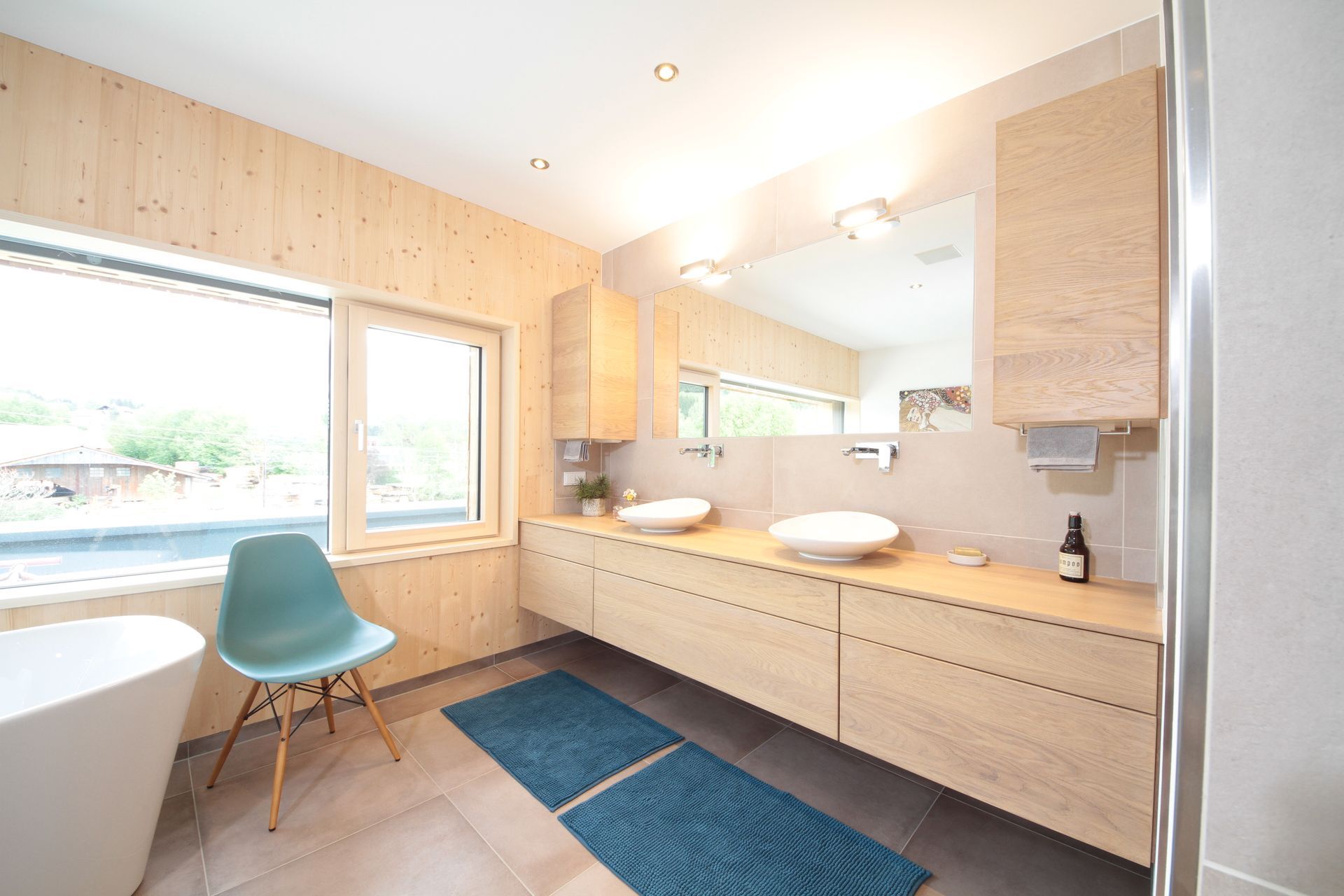 Helles, modernes Badezimmer mit Holzschränken, großem Spiegel, Fenster, Badewanne und blauem Stuhl