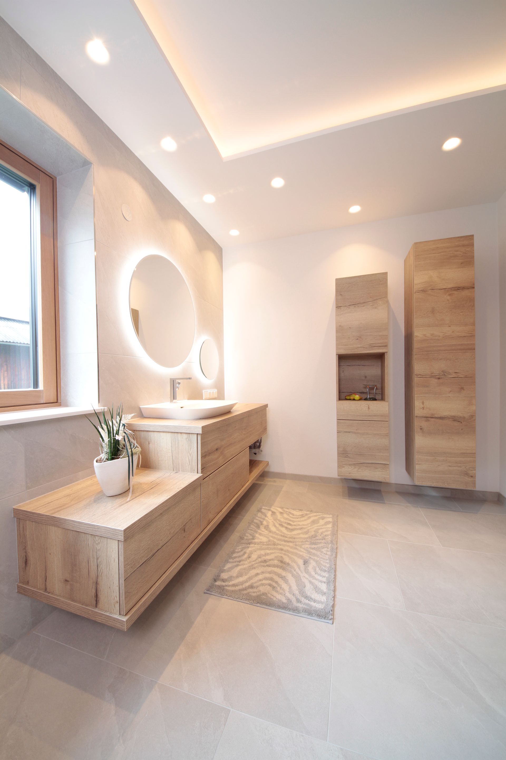 Helles, modernes Badezimmer mit Waschtisch aus Holz, rundem Spiegel, geflochtener Bank und zwei hohen Aufbewahrungsschränken.