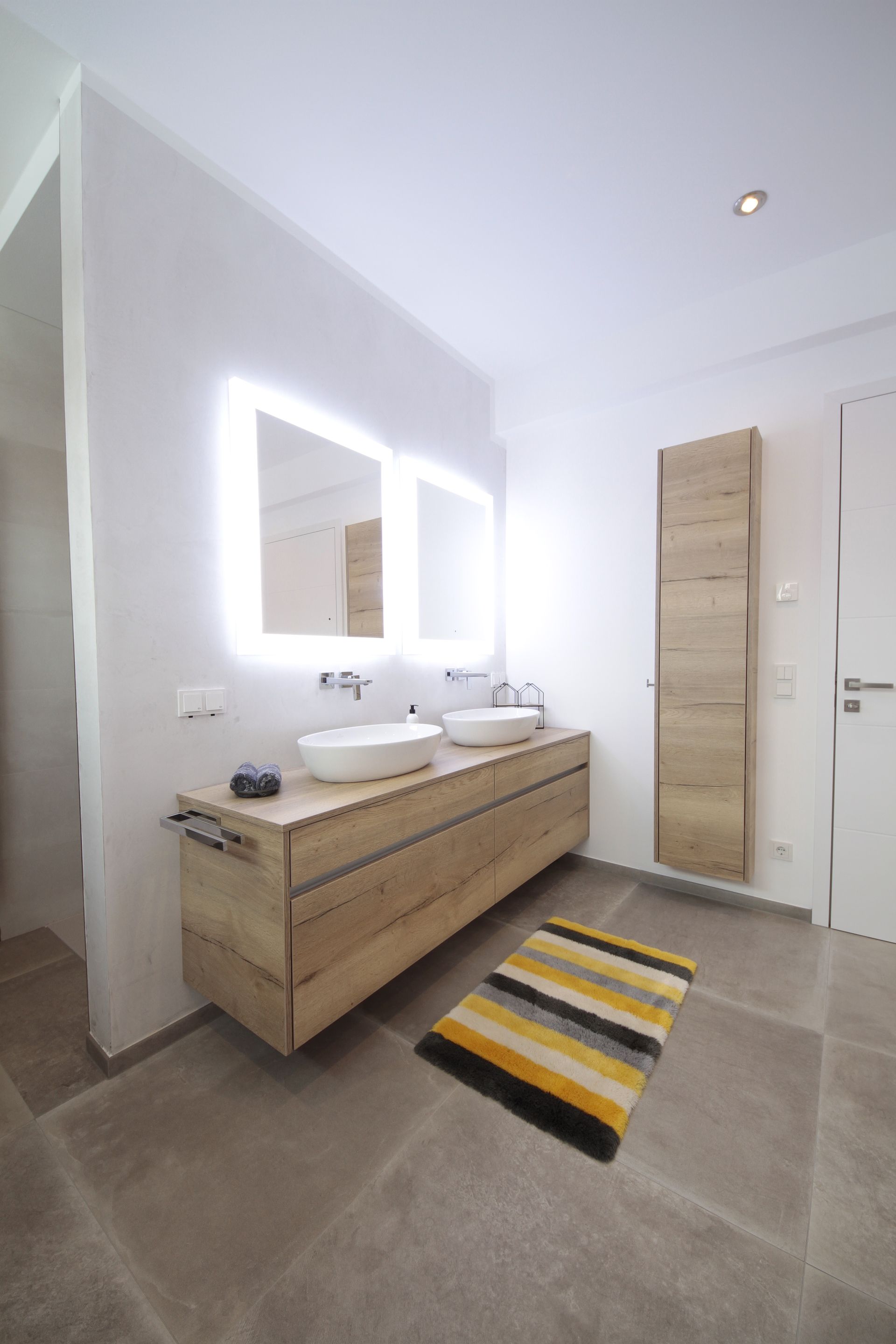 Modernes Badezimmer mit Doppelwaschtisch aus Holz, beleuchtetem Spiegel, Handtuchheizkörper und gestreiftem Teppich.