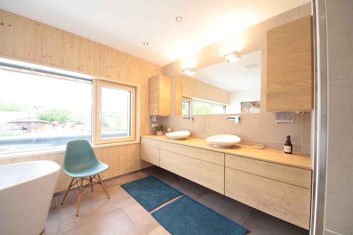 Helles, modernes Badezimmer mit Holzschränken, großem Spiegel, blauem Teppich und Stuhl am Fenster
