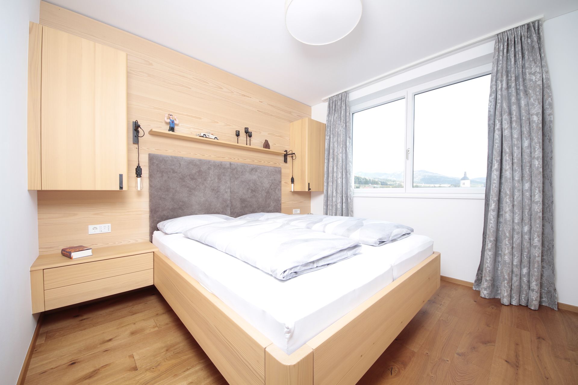 Helles Schlafzimmer mit Doppelbett, Holzboden, Kleiderschrank, Nachttisch und großem Fenster mit grauen Vorhängen.
