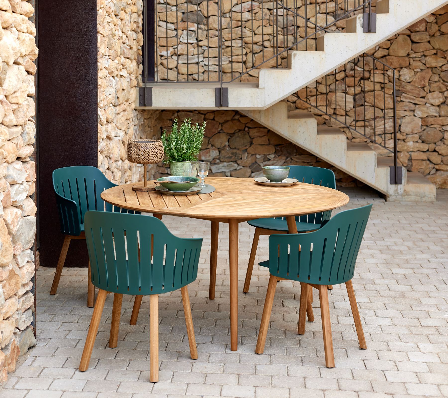 Terrasse im Freien mit rundem Holztisch und türkisfarbenen Stühlen neben einer Steinmauer und einer Treppe