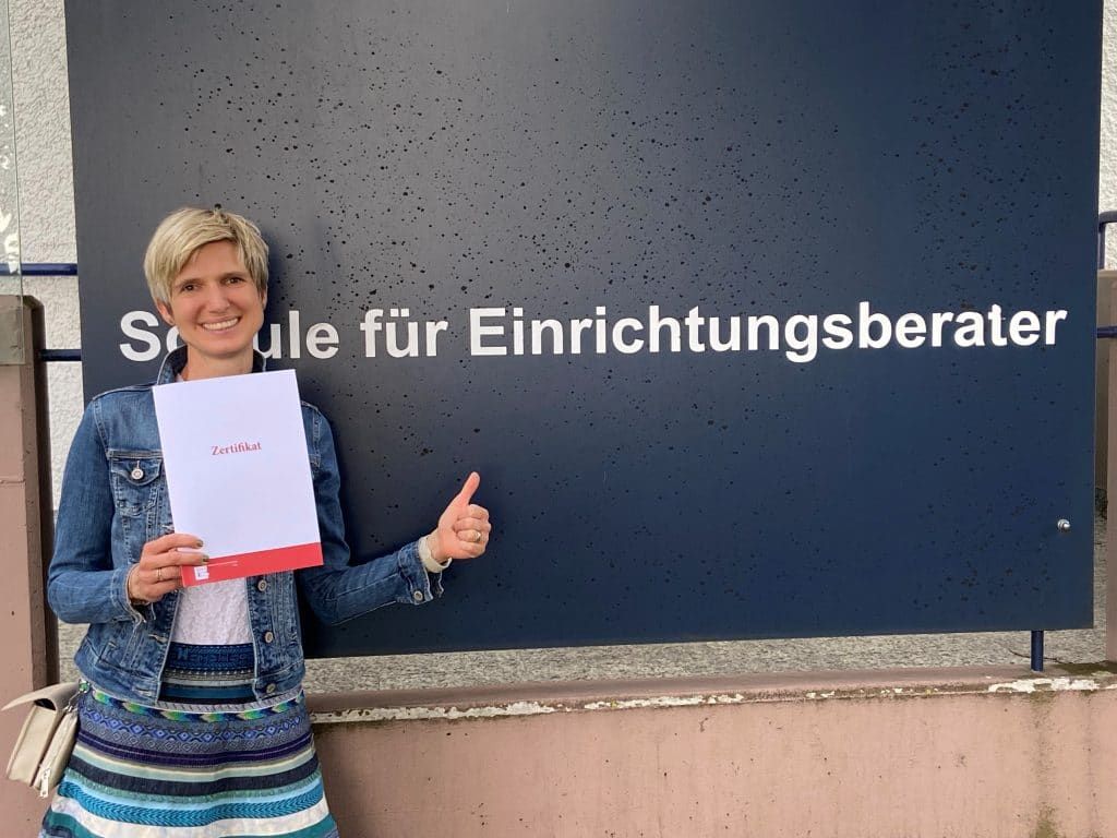 Frau zeigt den Daumen nach oben vor einem Schild mit der Aufschrift „Schule für Einrichtungsberater“.