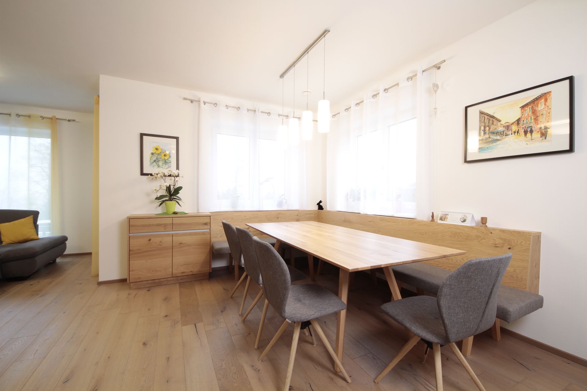 Helles Esszimmer mit Holztisch, grauen Stühlen, großen Fenstern und einem Sideboard mit Pflanzen und gerahmten Kunstwerken