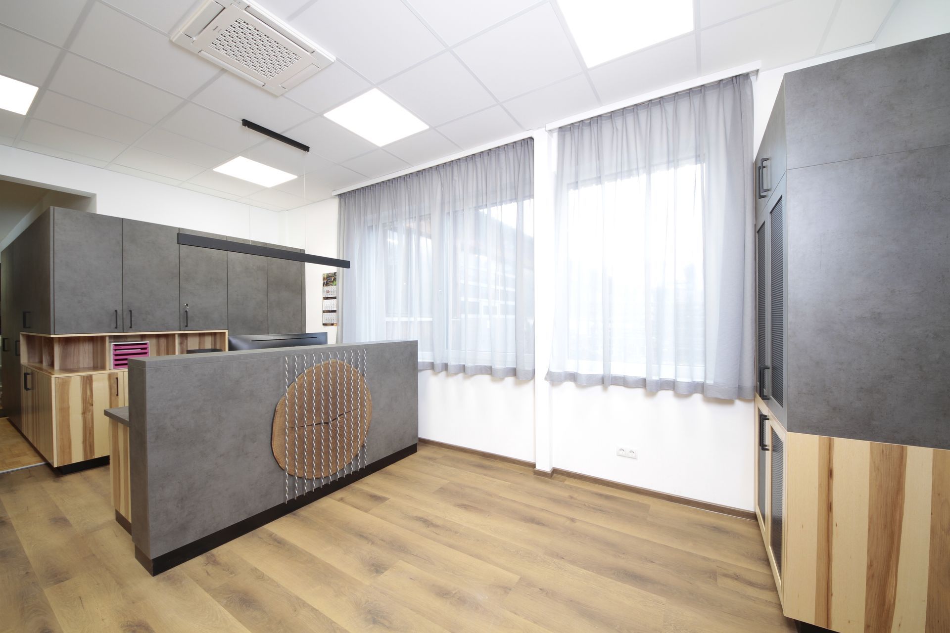 Modernes Bürointerieur mit Holzböden, grauen Theken und großen Fenstern mit transparenten Vorhängen.