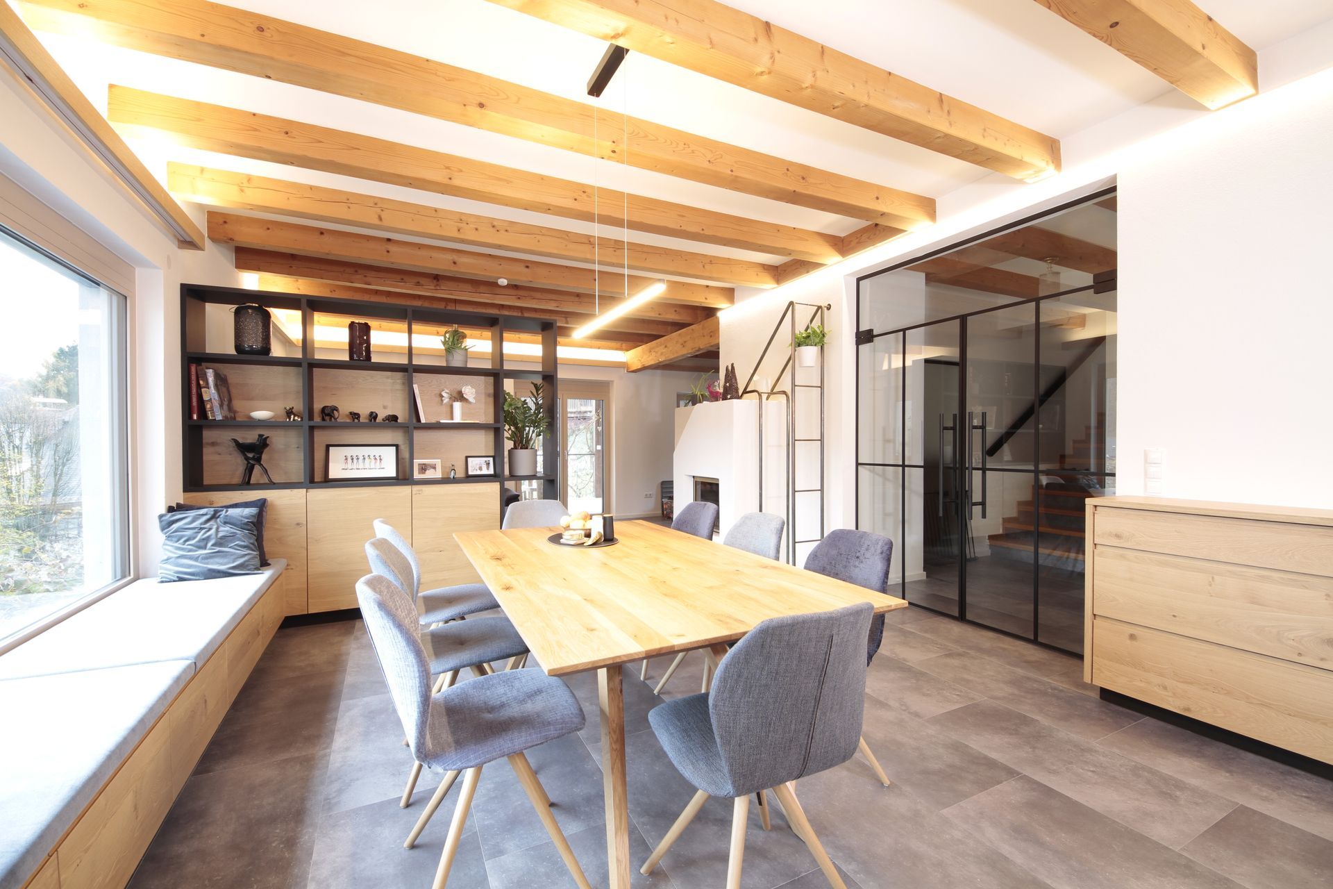 Helles Esszimmer mit Holzbalken, langem Tisch, grauen Stühlen, offenen Regalen und Glastüren.