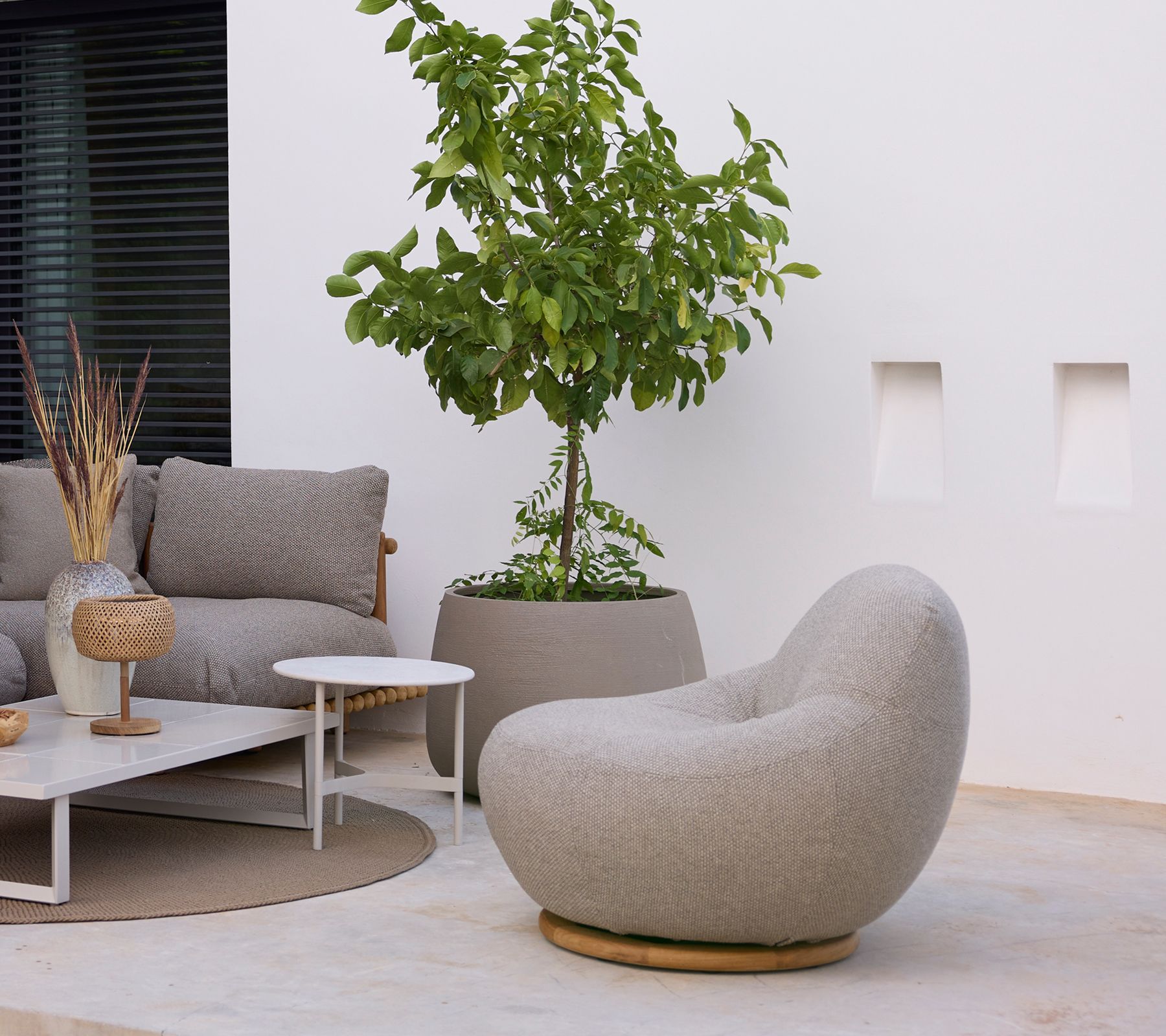 Moderne Terrasse mit grauen, strukturierten Stühlen, weißen Tischen, einem Topfbaum und einer minimalistischen weißen Wand