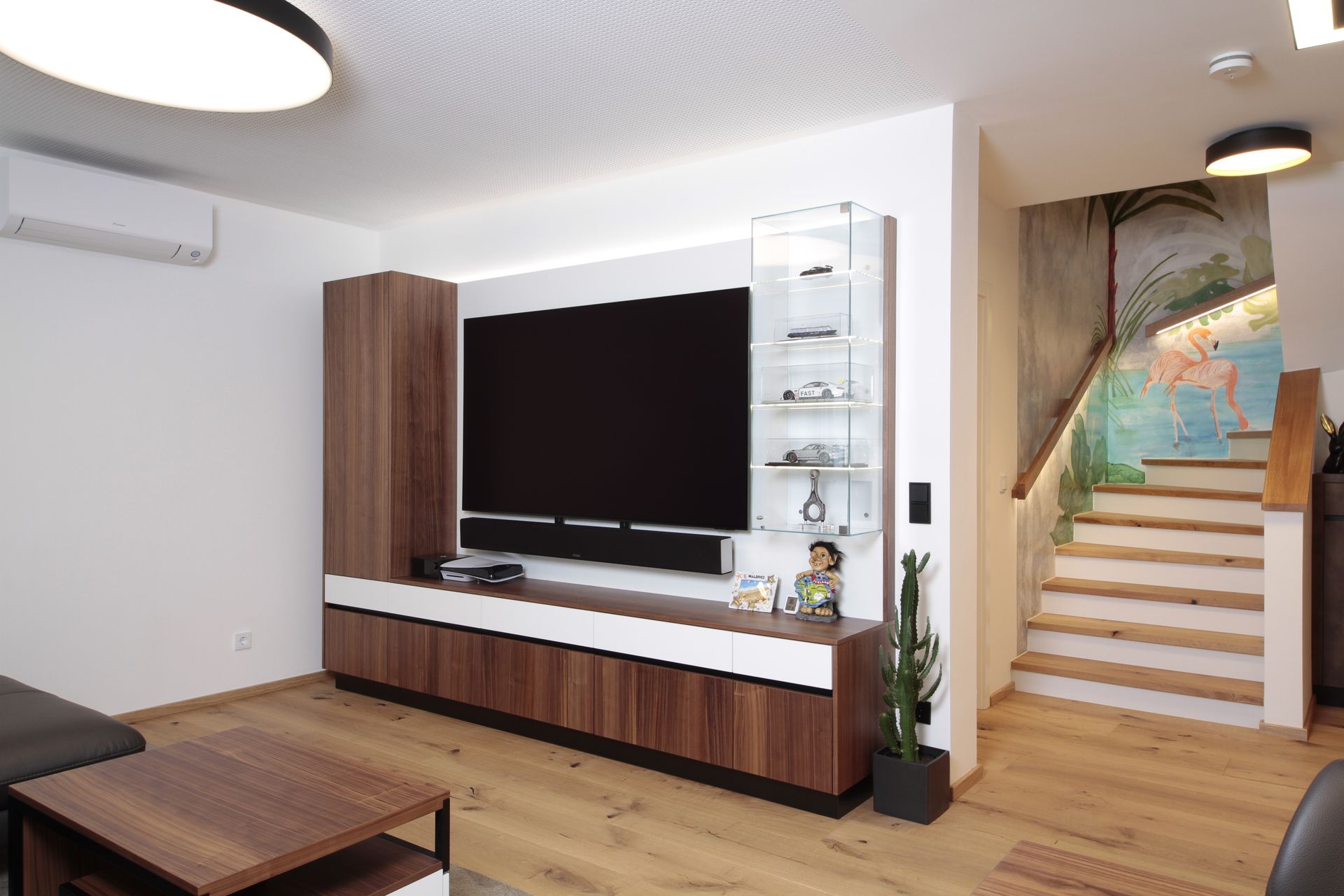 Modernes Wohnzimmer mit großem Fernseher, Holzboden und Treppe neben einem farbenfrohen Wandbild