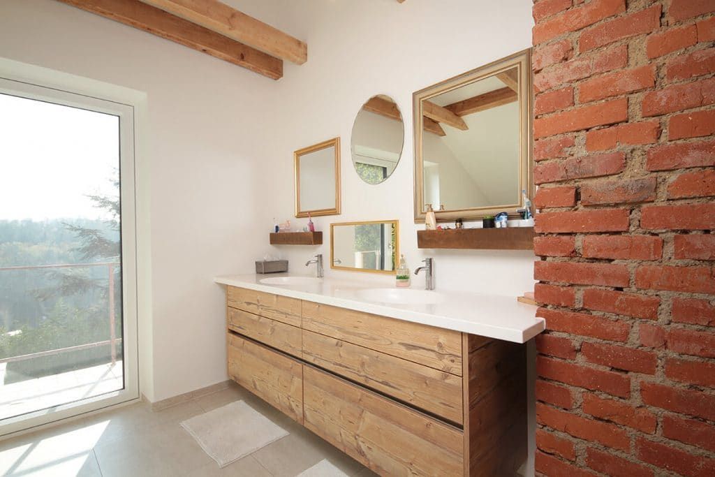 Modernes Badezimmer mit Waschtisch aus Holz, weißer Arbeitsplatte, rundem Spiegel und freiliegender Ziegelwand