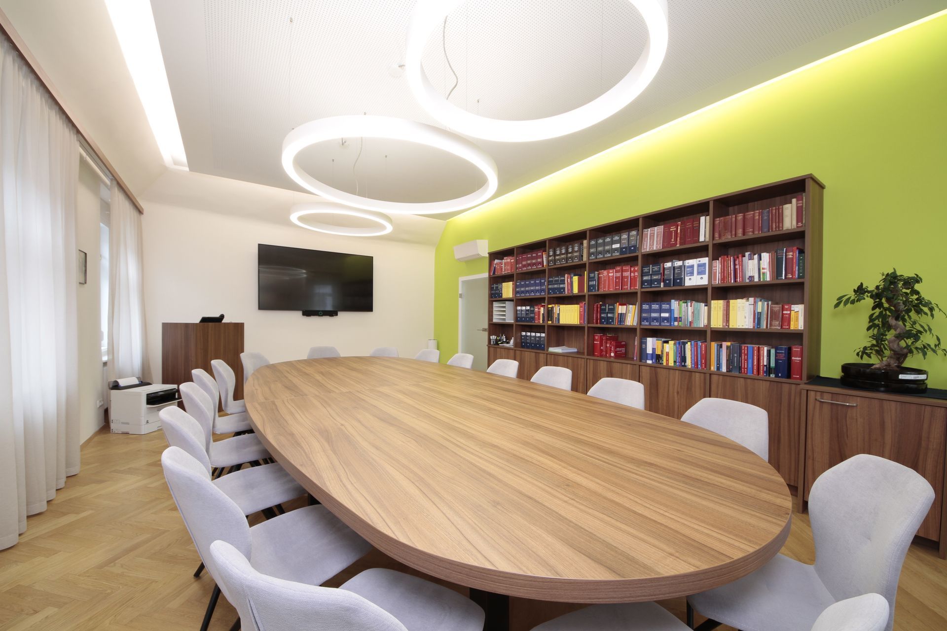 Moderner Konferenzraum mit ovalem Holztisch, grauen Stühlen, Wandfernseher, Bücherregalen und grüner Akzentwand