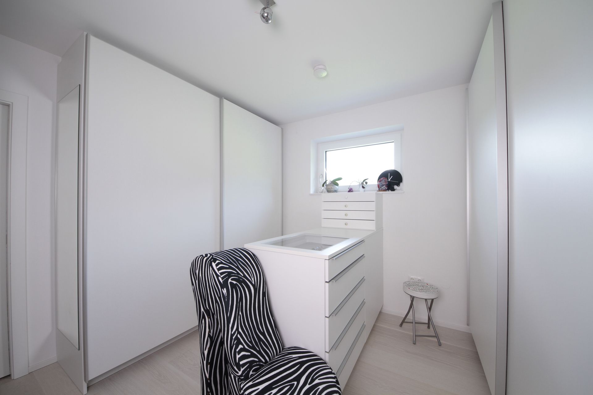 Helles, weißes Schlafzimmer mit Kommode, Kleiderschrank, gemustertem Stuhl, kleinem Fenster und Hocker.