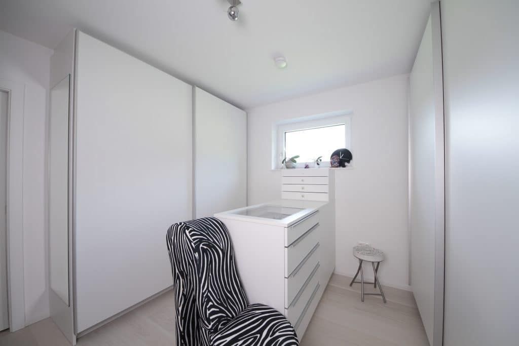 Helles, weißes Schlafzimmer mit Kommode, Stuhl mit Zebramuster, kleinem Fenster und Tritthocker.
