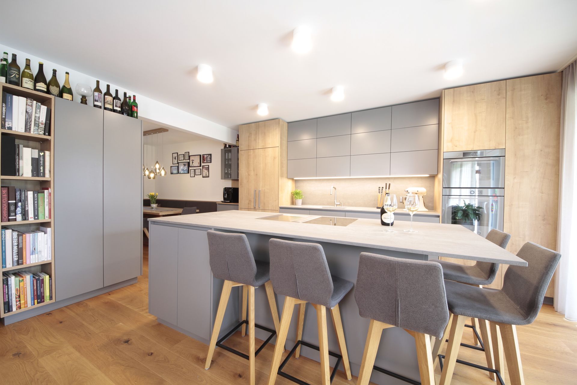 Moderne Küche mit grauer Kochinsel, Barhockern, Holzboden und eingebautem Kühlschrank neben einem Weinregal.