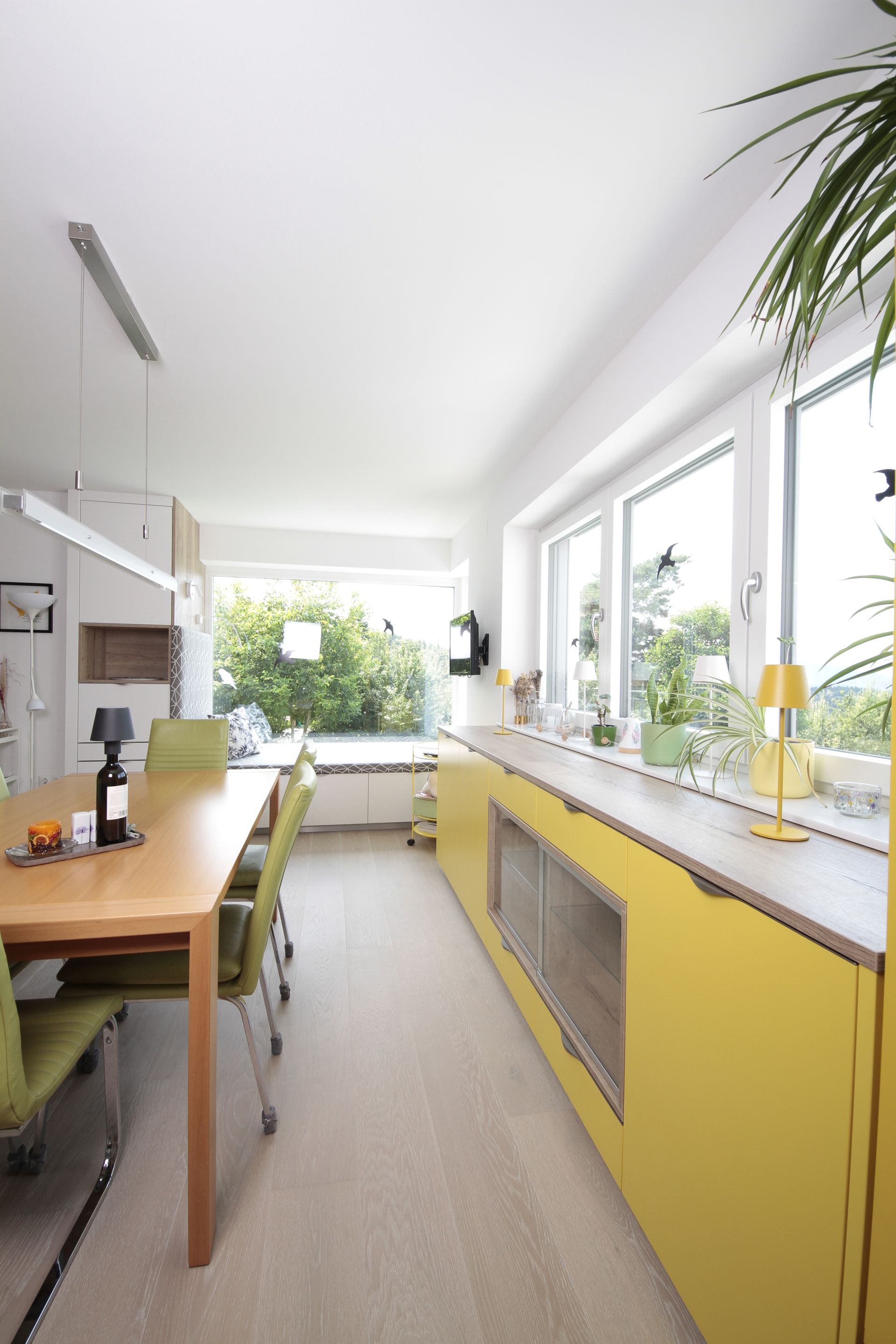Helle Küche mit gelben Schränken, Esstisch aus Holz und großen Fenstern mit Blick ins Grüne.