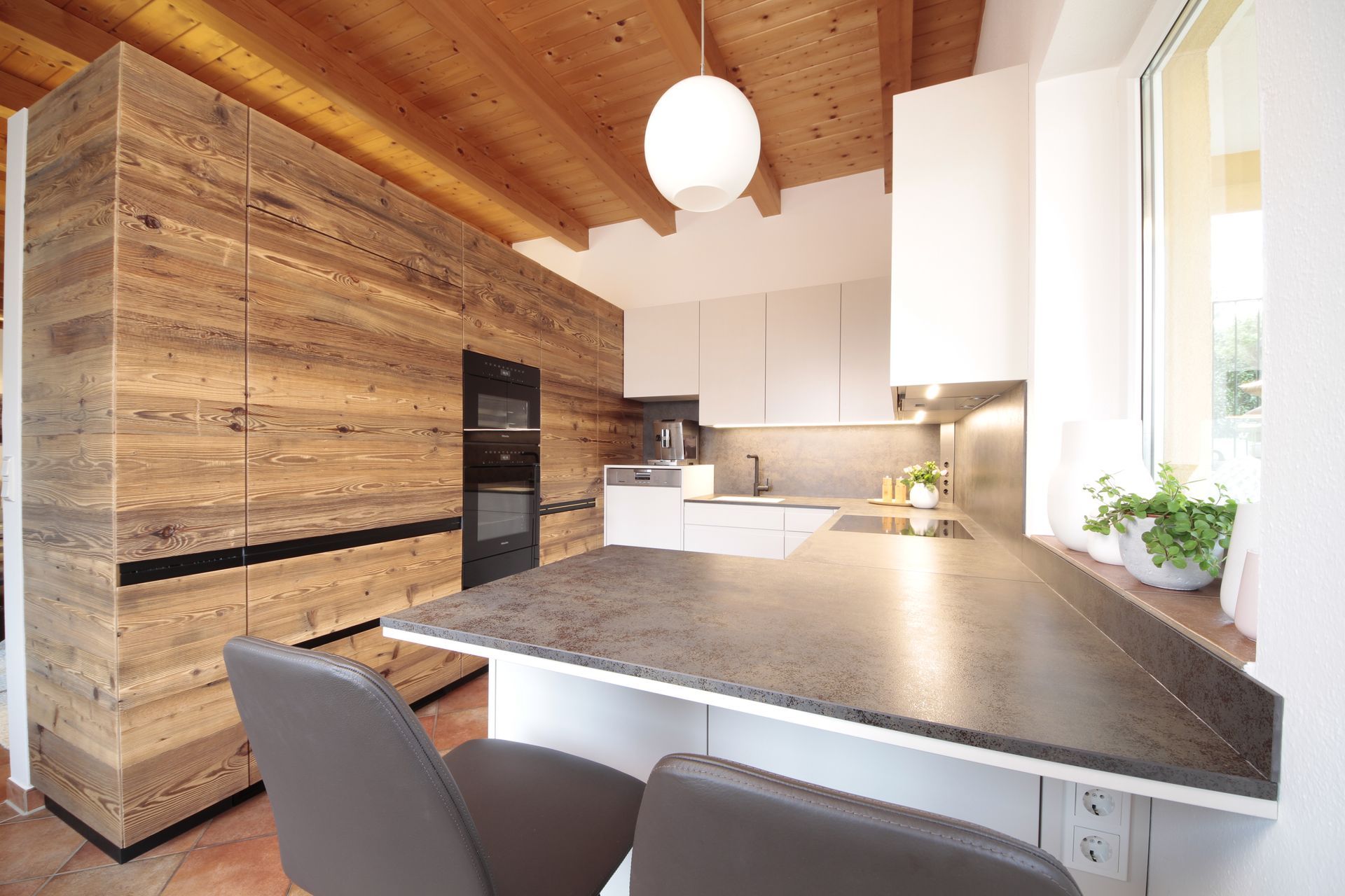 Moderne Küche mit Holzdecke, hellen Schränken und einem großen Fenster neben einem grauen Tisch.