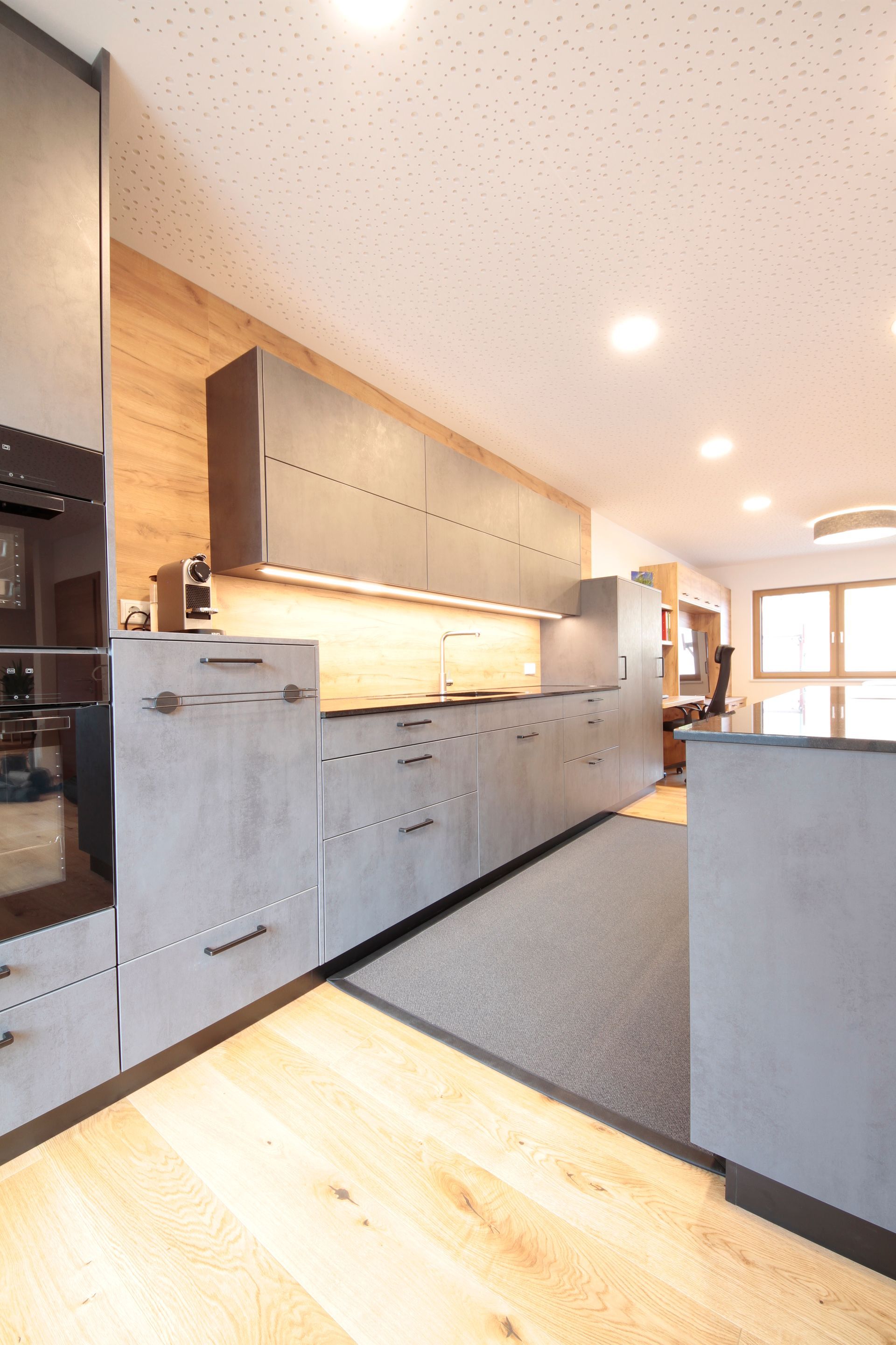 Moderne Küche mit grauen Schränken, Holzboden und heller Deckenbeleuchtung, vom Eingangsbereich aus gesehen
