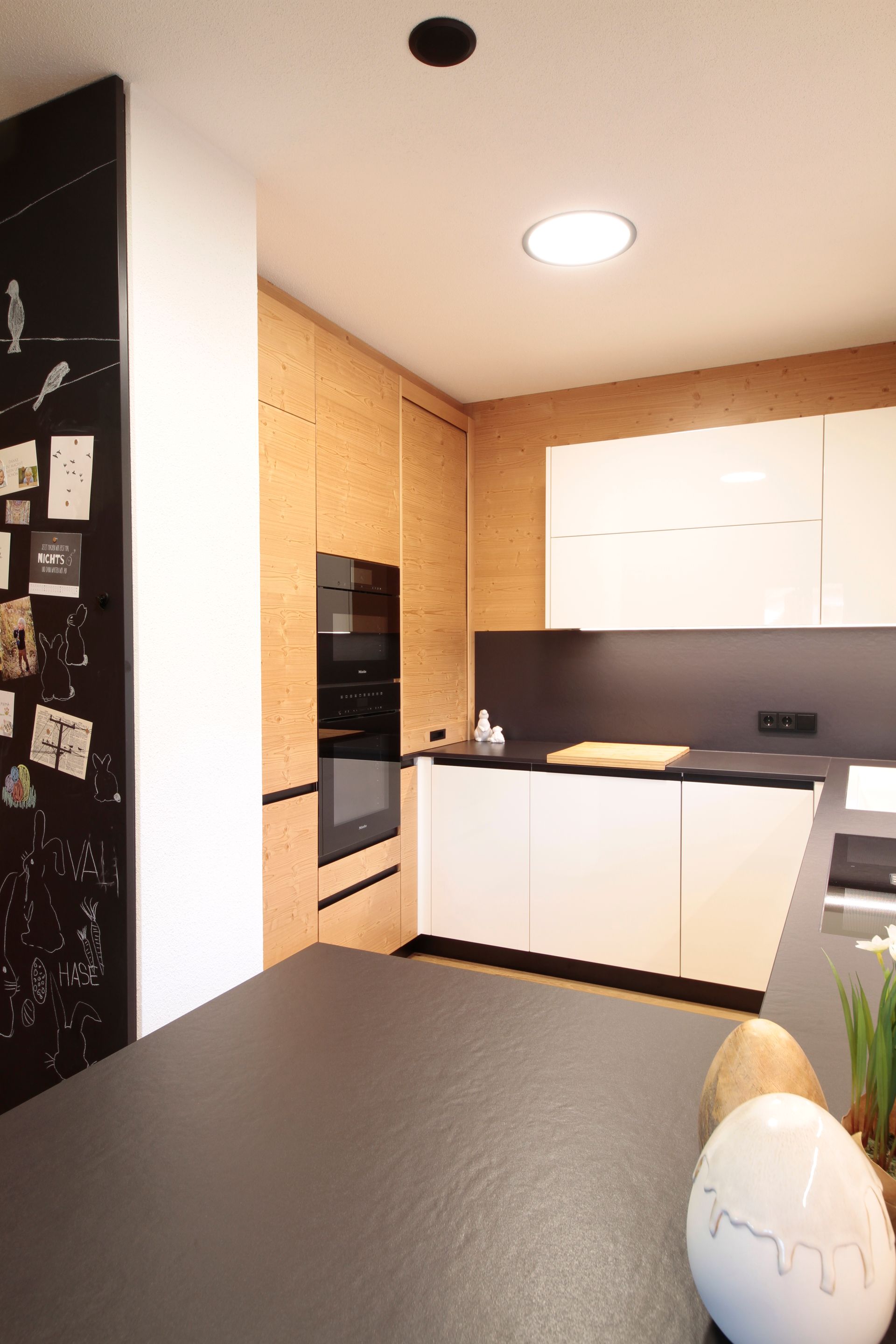 Moderne Küche mit schwarzer Arbeitsplatte, weißen Schränken, eingebautem Backofen und warmen, holzvertäfelten Wänden.