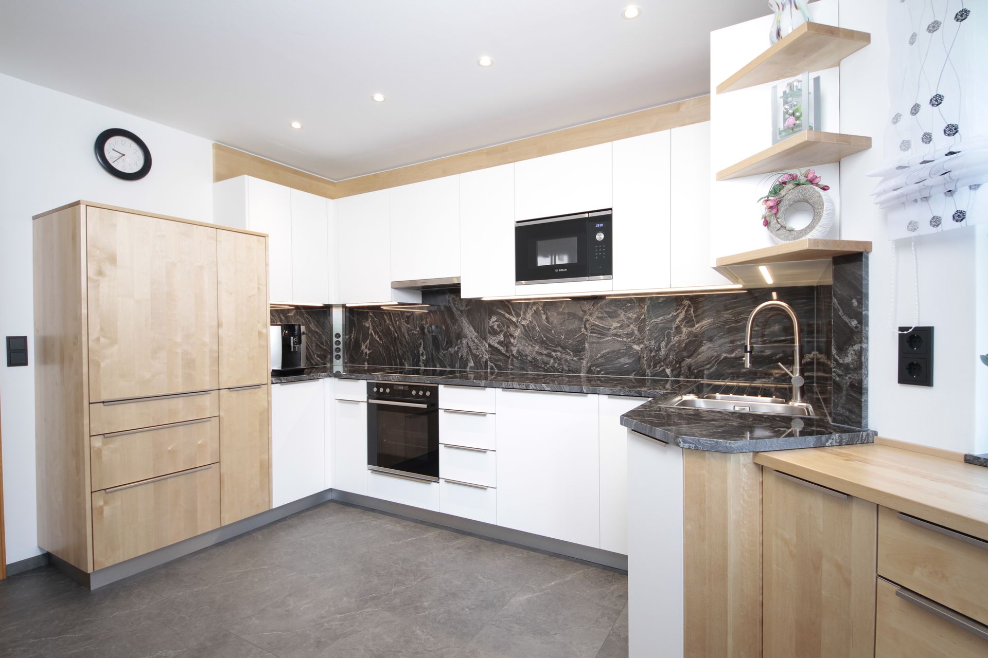 Moderne Küche mit weißen Schränken, schwarzen Arbeitsplatten, Holzelementen sowie Kühlschrank, Backofen und Mikrowelle.