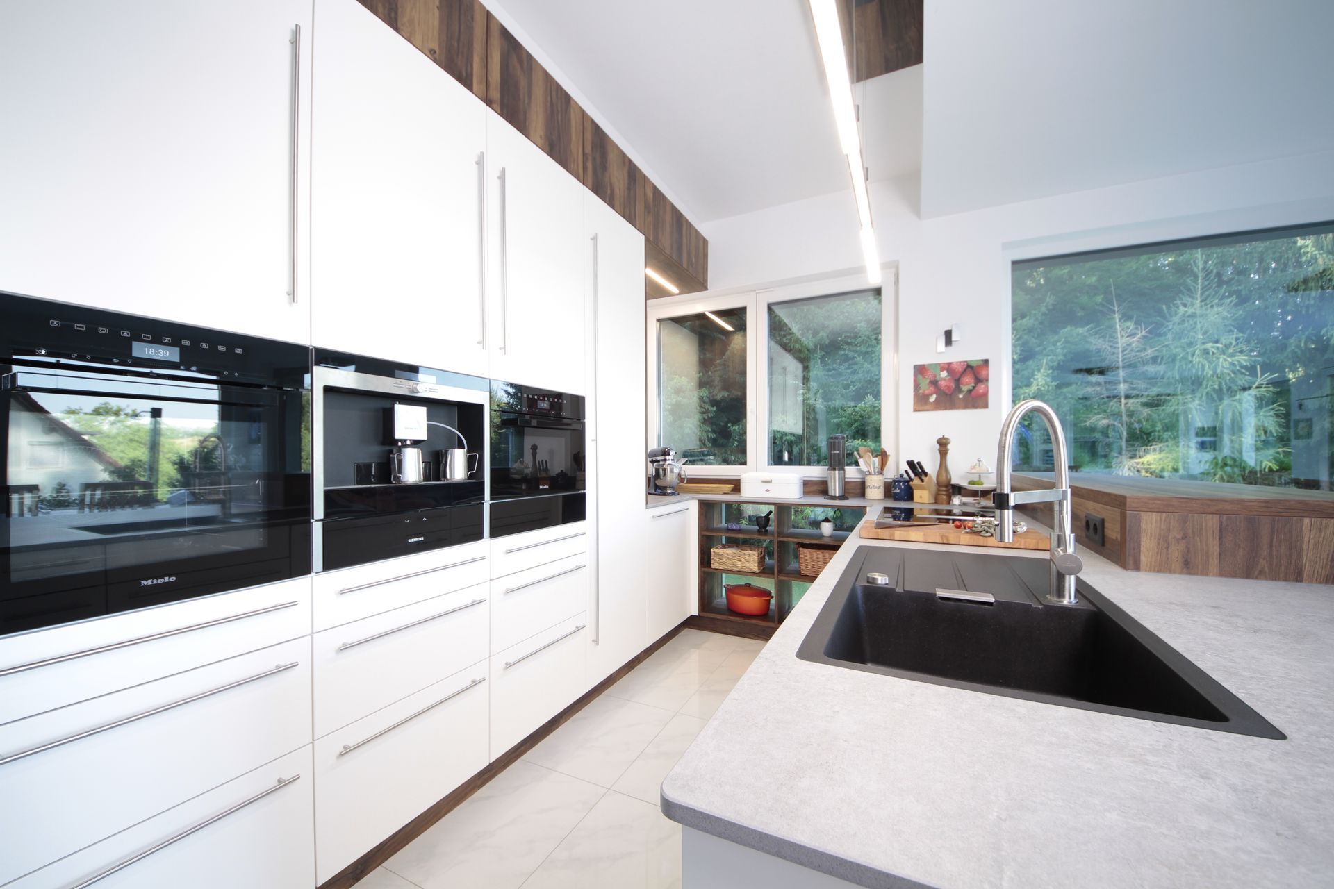 Moderne weiße Küche mit Einbaubacköfen, großer Kücheninsel mit Spülbecken und breiten Fenstern mit Blick ins Grüne