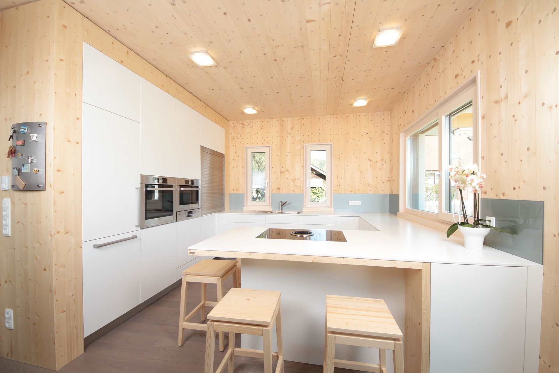 Helle, moderne Küche mit weißen Schränken, Holzdecke, Kochinsel und Barhockern in der Nähe großer Fenster.
