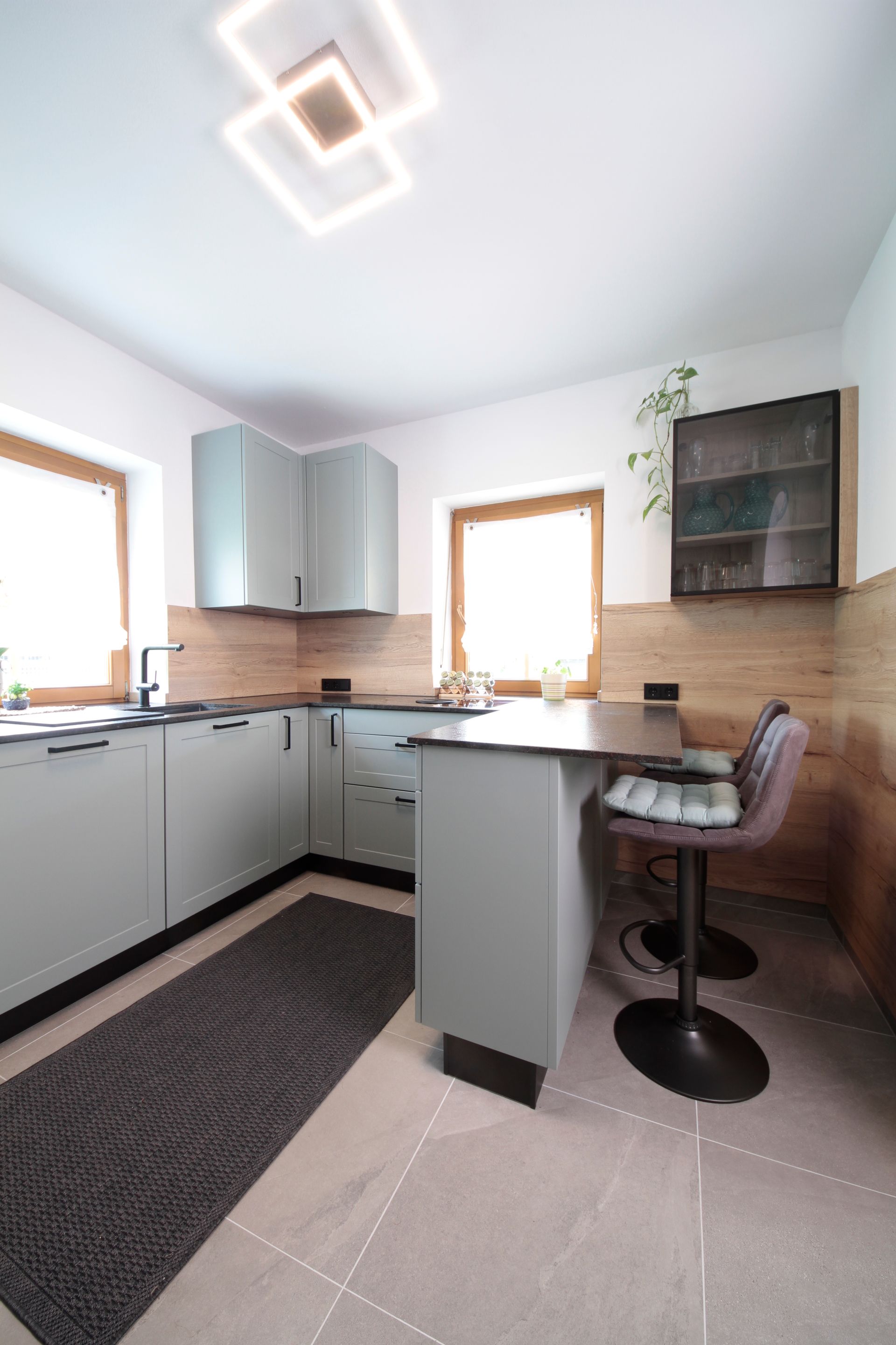 Moderne Küche mit grauen Schränken, Holzelementen, einer Kochinsel und einem Barhocker an der Theke.