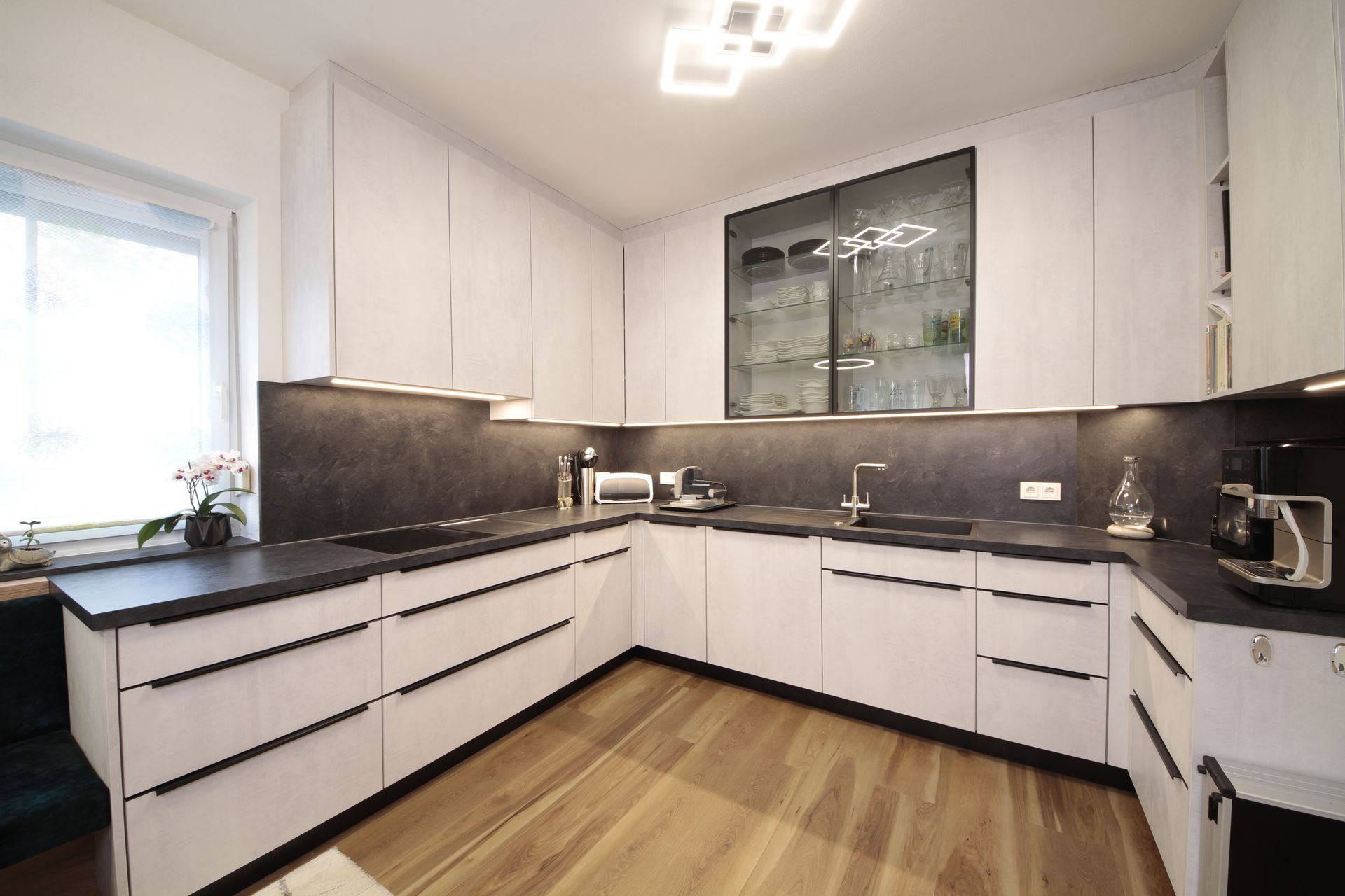 Moderne weiße Küche mit schwarzen Arbeitsplatten, Holzboden und Schranktüren aus Glas.