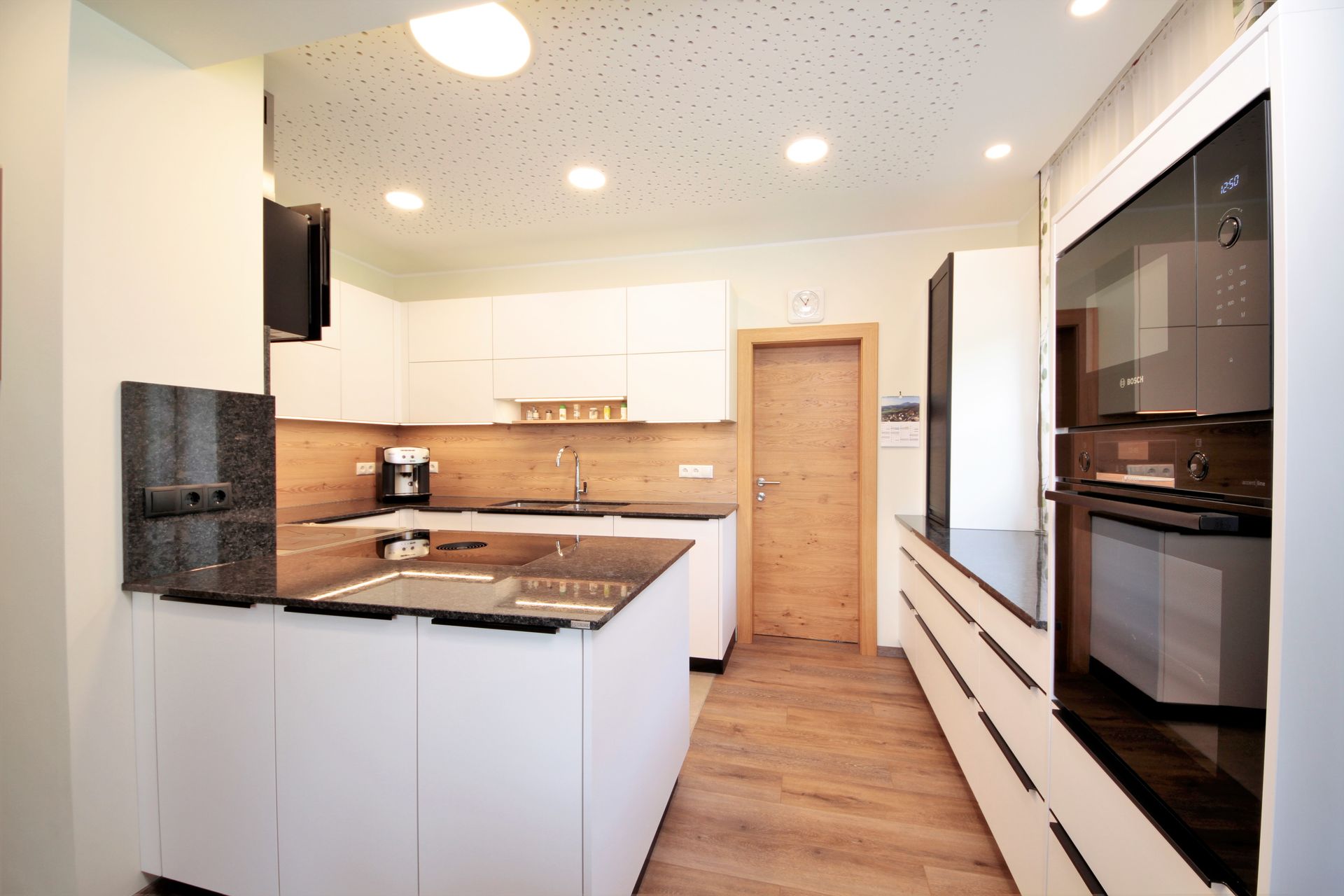 Moderne weiße Küche mit schwarzen Arbeitsplatten, Holzboden und eingebauten Edelstahlgeräten