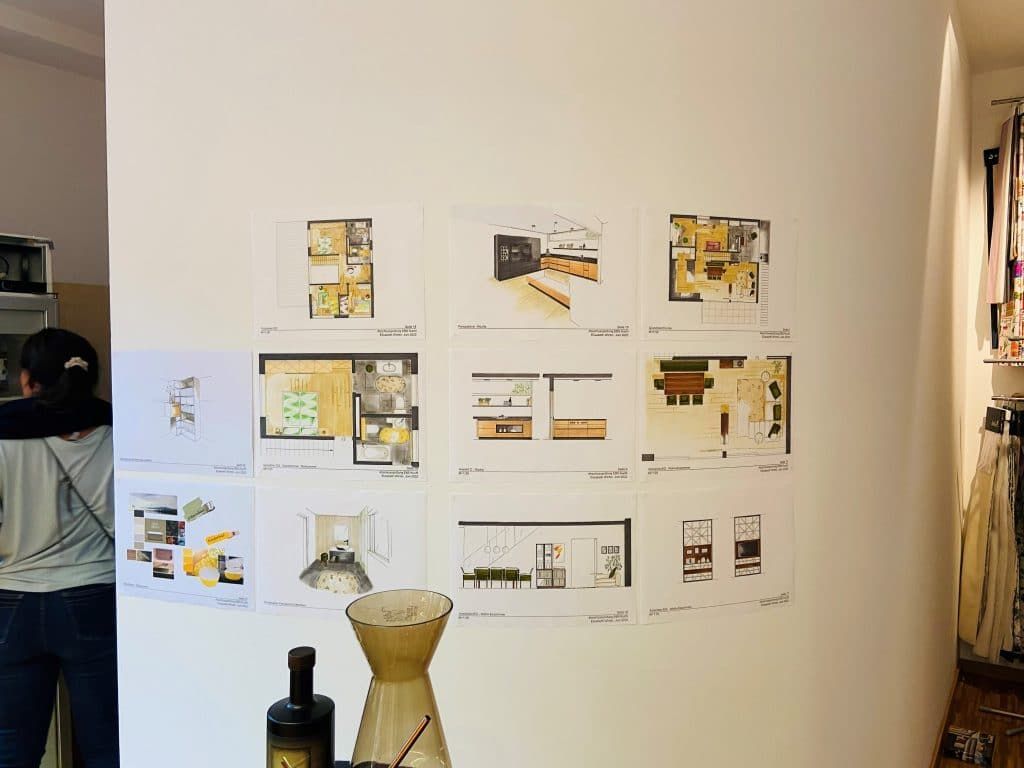 Eine Wand mit einem Raster aus Designskizzen und Fotos über einem Tisch mit einer Flasche und einer Vase.