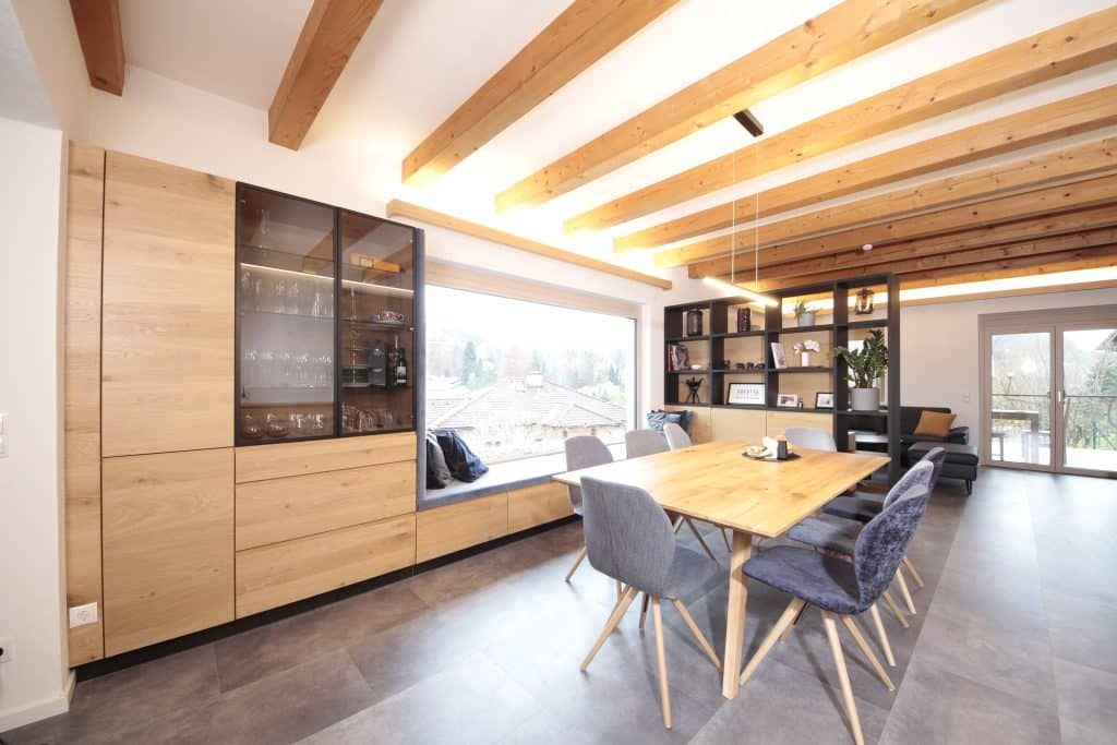 Modernes Esszimmer mit Holzbalkendecke, langem Tisch, Stühlen und Einbauschränken an großen Fenstern