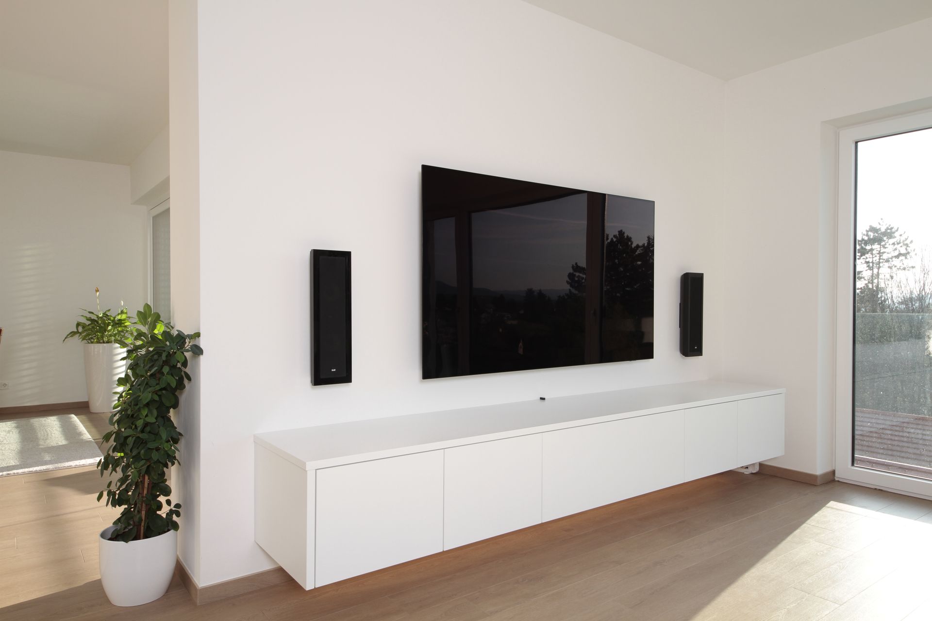 Modernes Wohnzimmer mit an der Wand montiertem Fernseher, Lautsprechern, weißem Mediaschrank, Pflanze und Glasschiebetür.