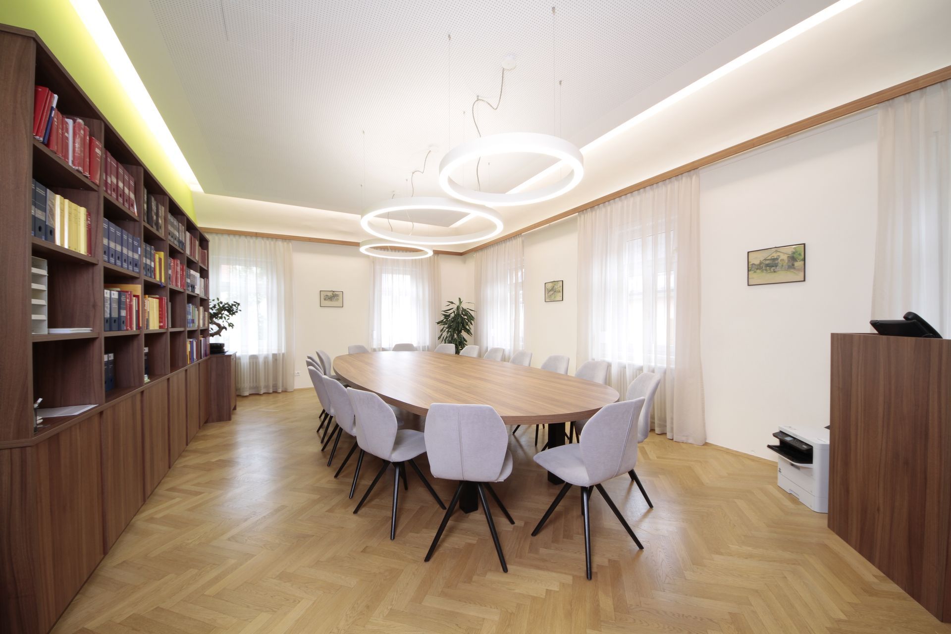 Heller Konferenzraum mit langem Tisch, grauen Stühlen, Bücherregalen und ringförmigen Deckenleuchten