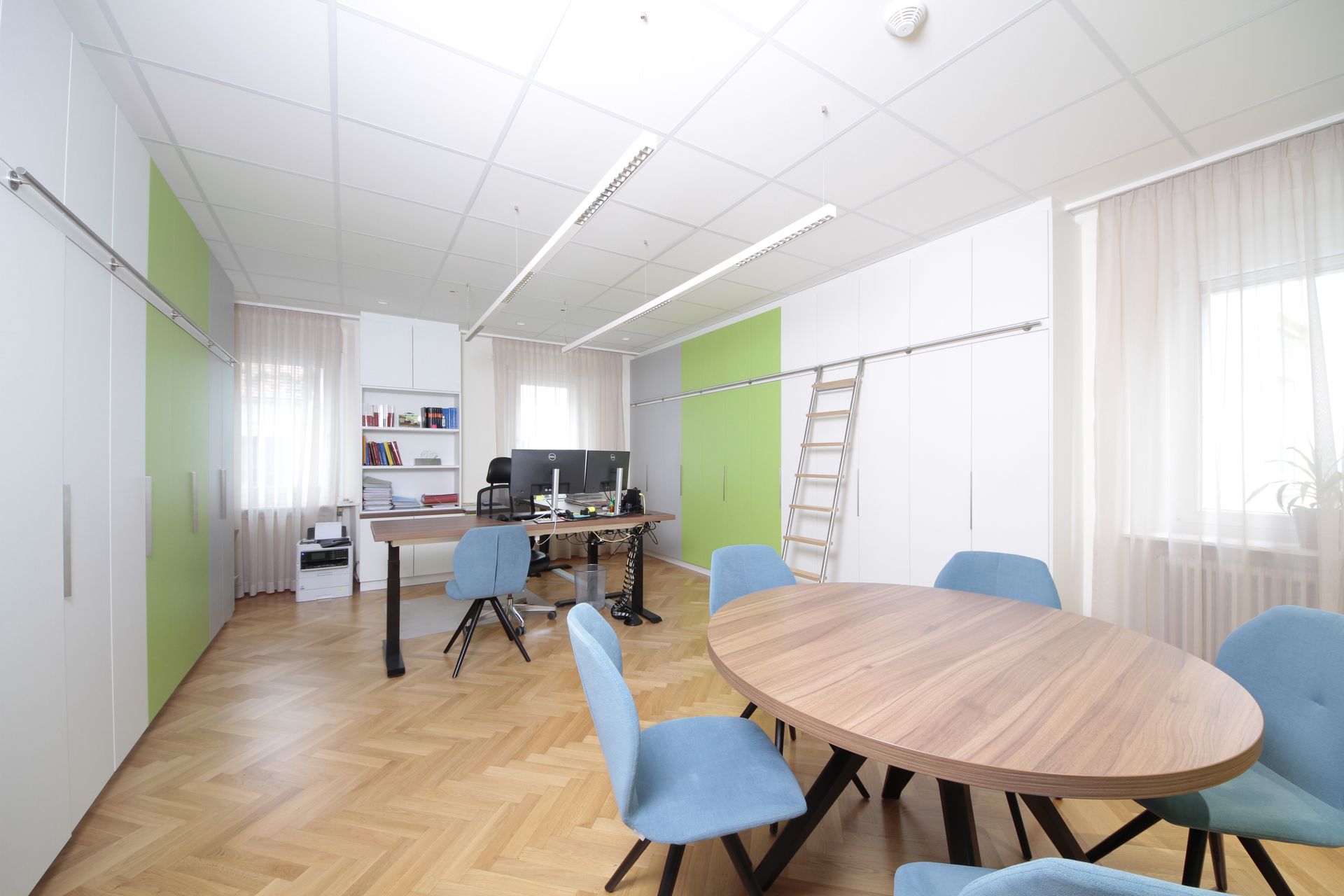 Helles Büro mit runden Holztischen, blauen Stühlen, grünen Wandpaneelen und großen Fenstern