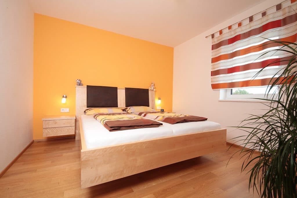 Modernes Schlafzimmer mit Holzbett, orangefarbener Akzentwand und gestreiften Fensterrollos.